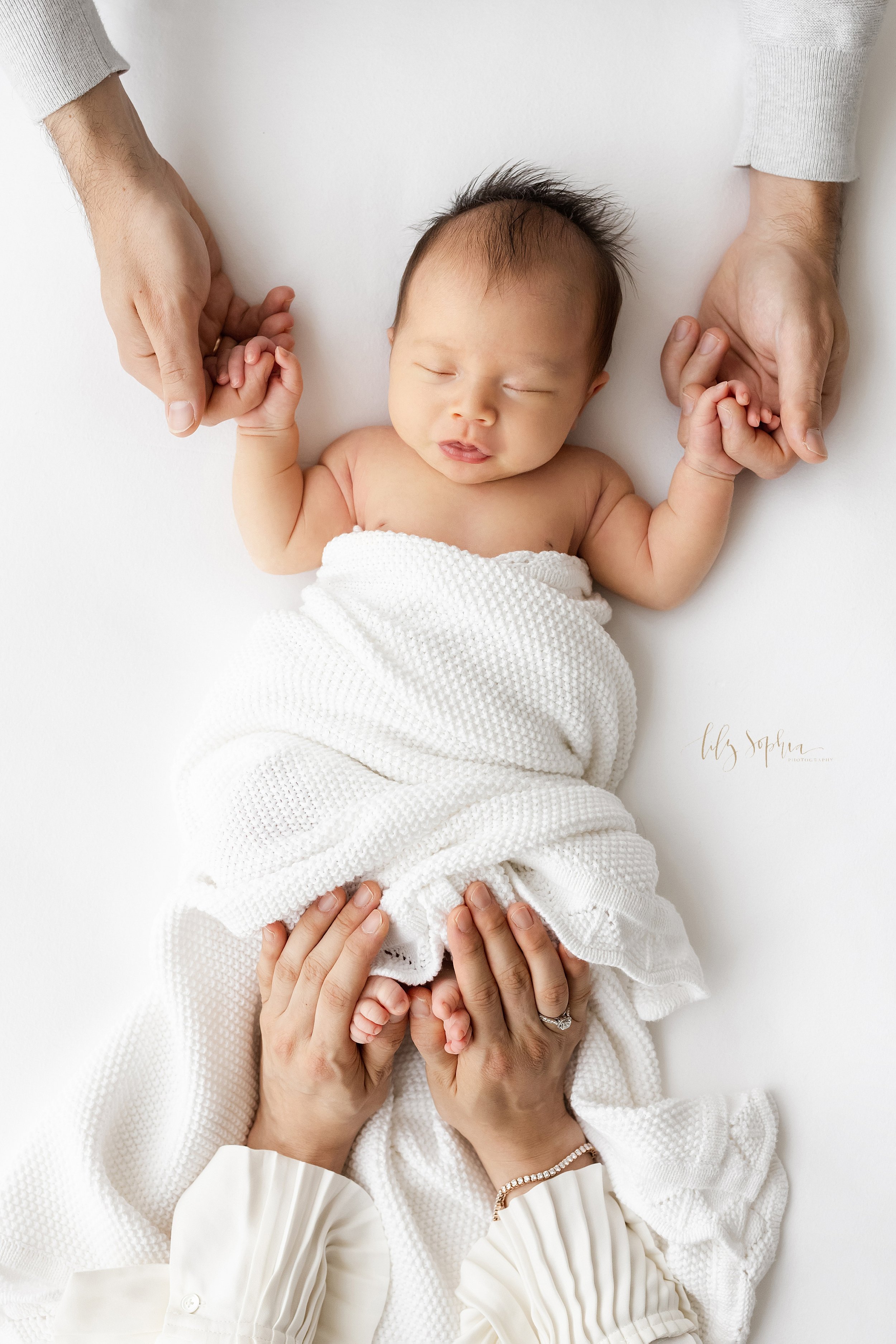  Newborn family photo of an infant Asian girl wrapped in a soft white blanket and sticking out her tongue as she holds her father’s fingers and her mother hold’s her daughter’s toes taken using natural light in a studio near Cummings in Atlanta. 