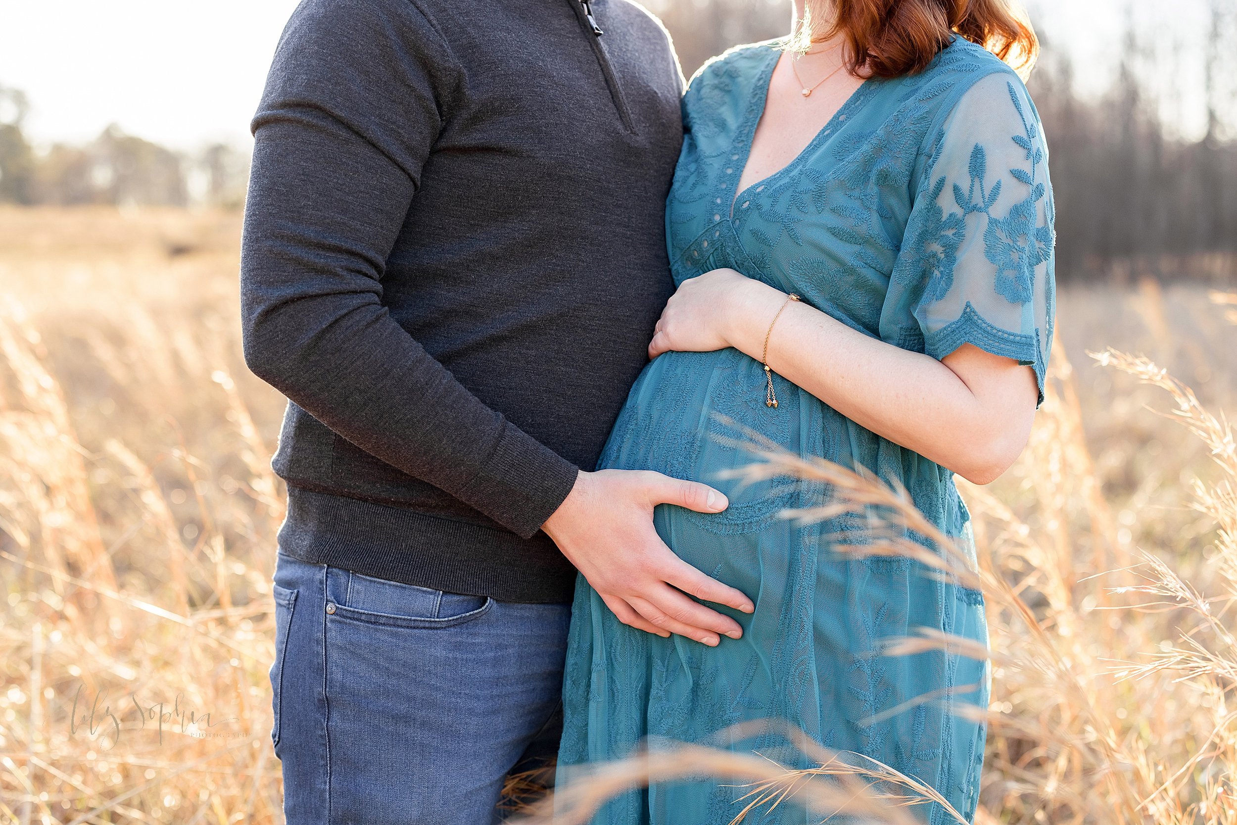  Maternity portrait of a pregnant mother resting her arm on the top of her belly as her husband holds their child in utero taken near Atlanta in a field during the fall season at sunset. 