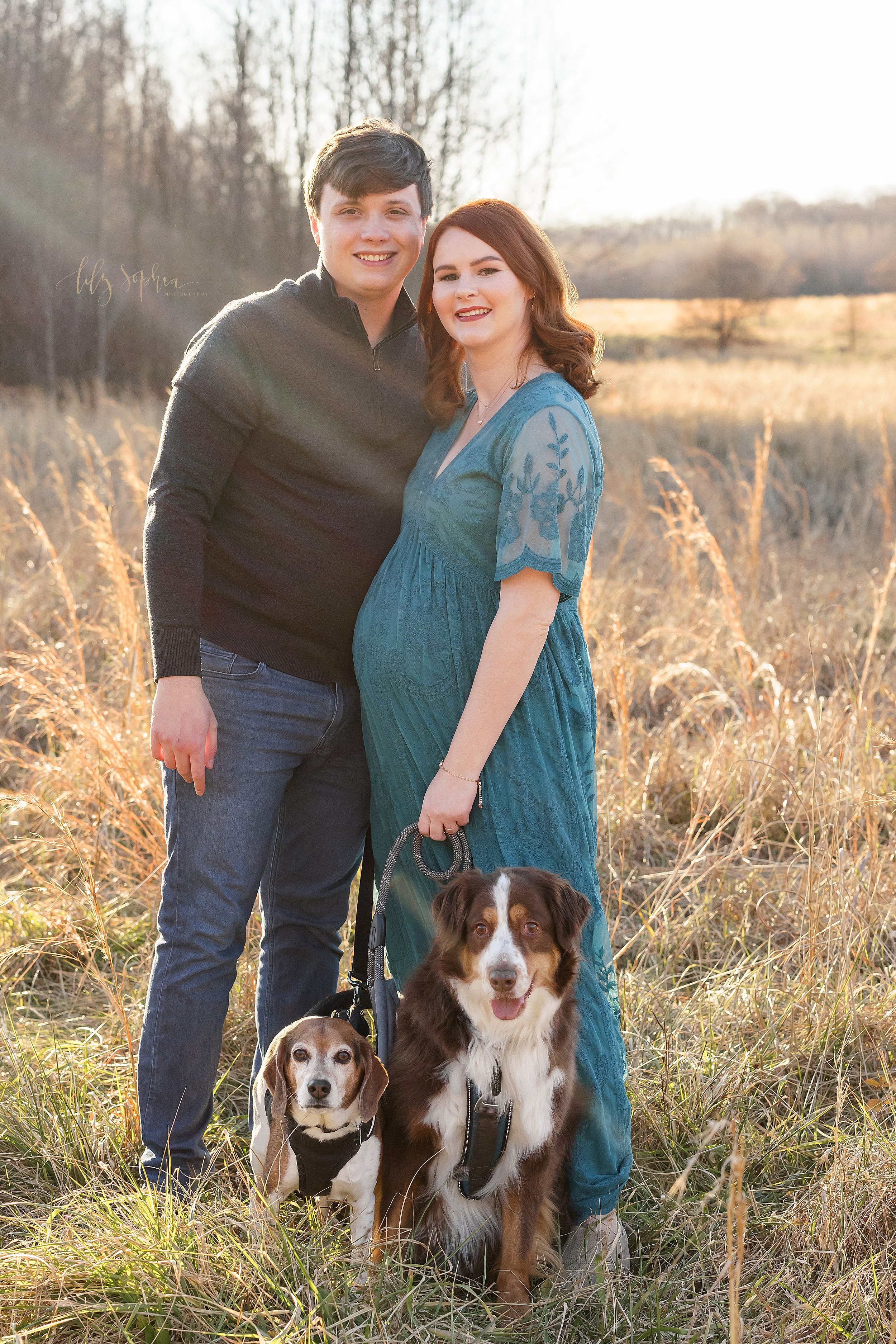  Maternity portrait of an expectant couple as they stand in a field at sunset during the winter season with their two dogs near Atlanta. 