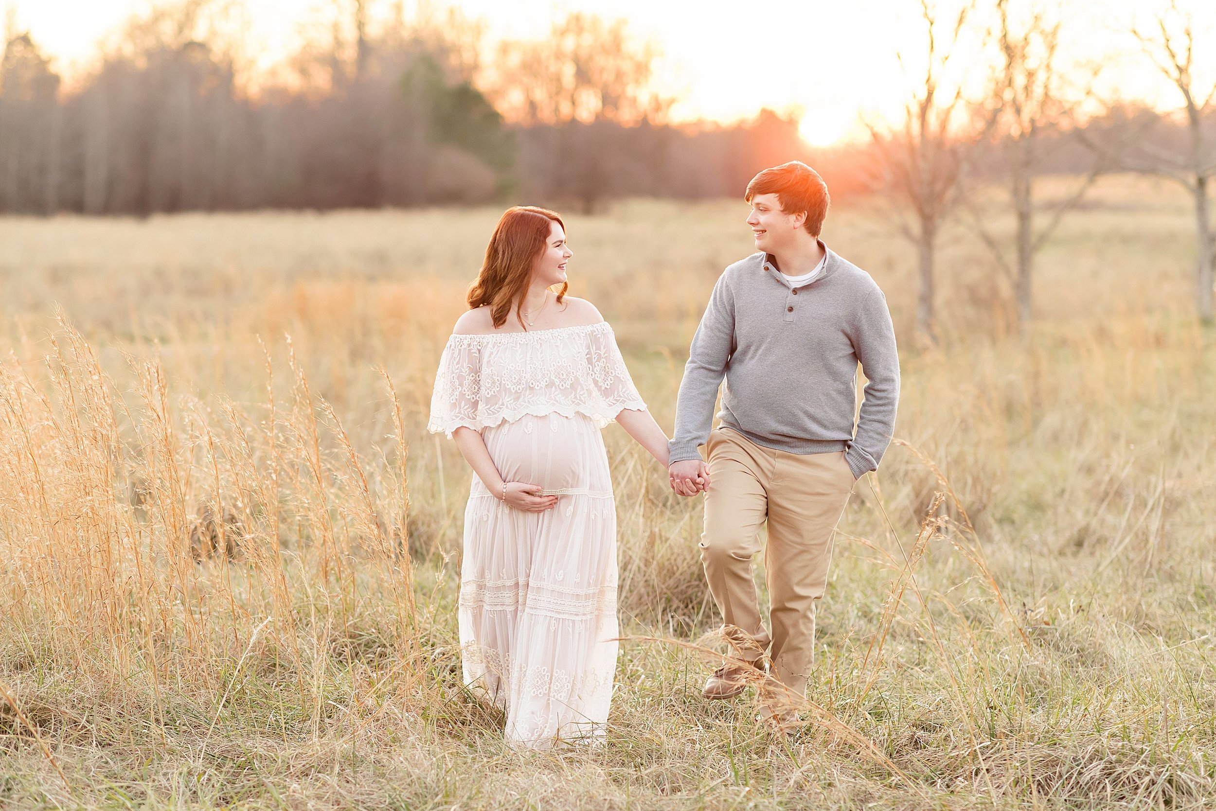  Maternity photo session of a pregnant red-haired mother as she walks hand-in-hand with her husband during autumn at sunset in a field near Atlanta. 