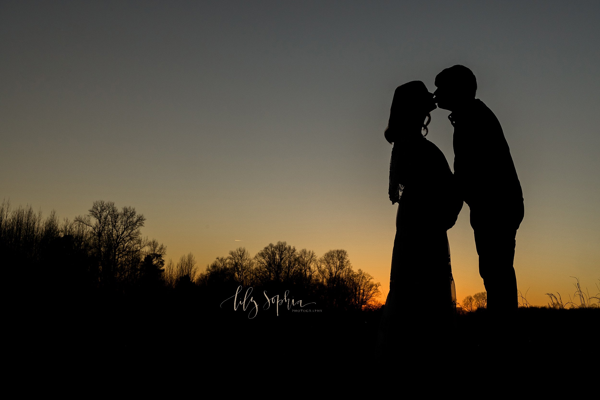  Maternity silhouetted portrait of an expectant mother kissing her husband as they stand atop a hill in a field at sunset near Atlanta, Georgia. 