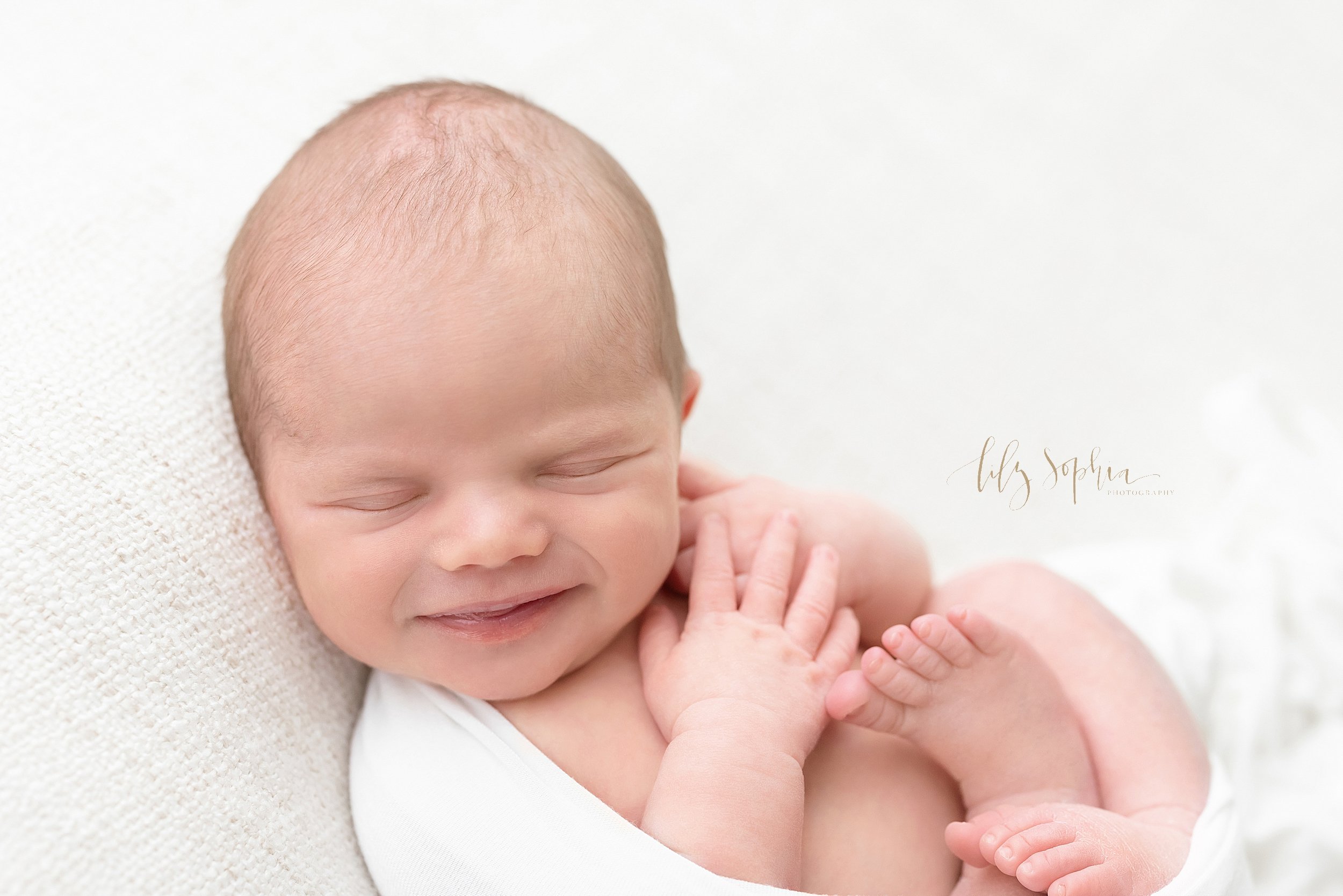  Newborn portrait of a smiling sleeping infant boy as he lies on his back wrapped in a stretchy swaddle taken in a natural light studio near Old Fourth Ward in Atlanta. 