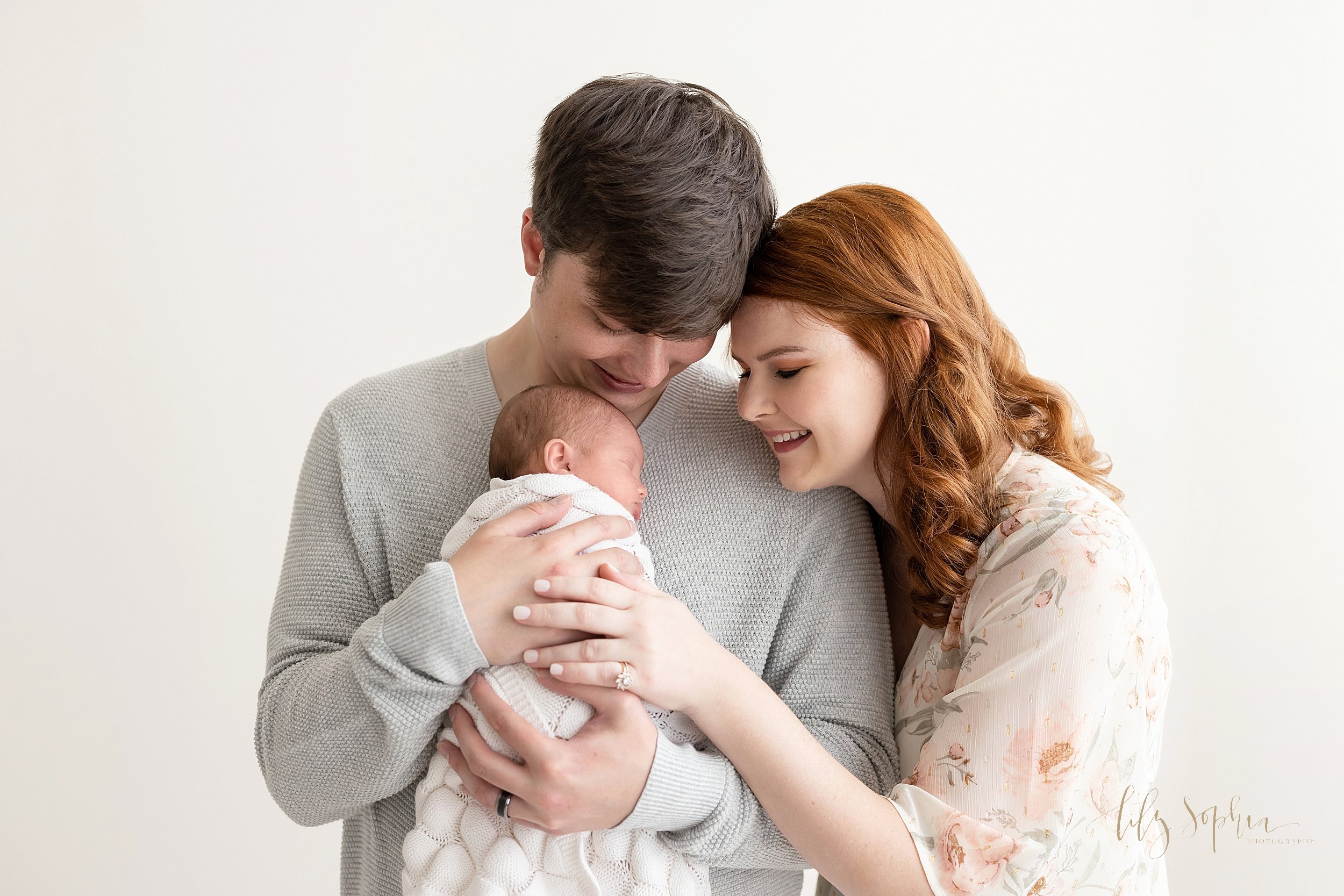  Family newborn portrait of a father holding his infant son against his chest as his wife leans her head against her husbands and looks over his shoulder at their little one taken in front of a window streaming natural light in a studio in Ponce City