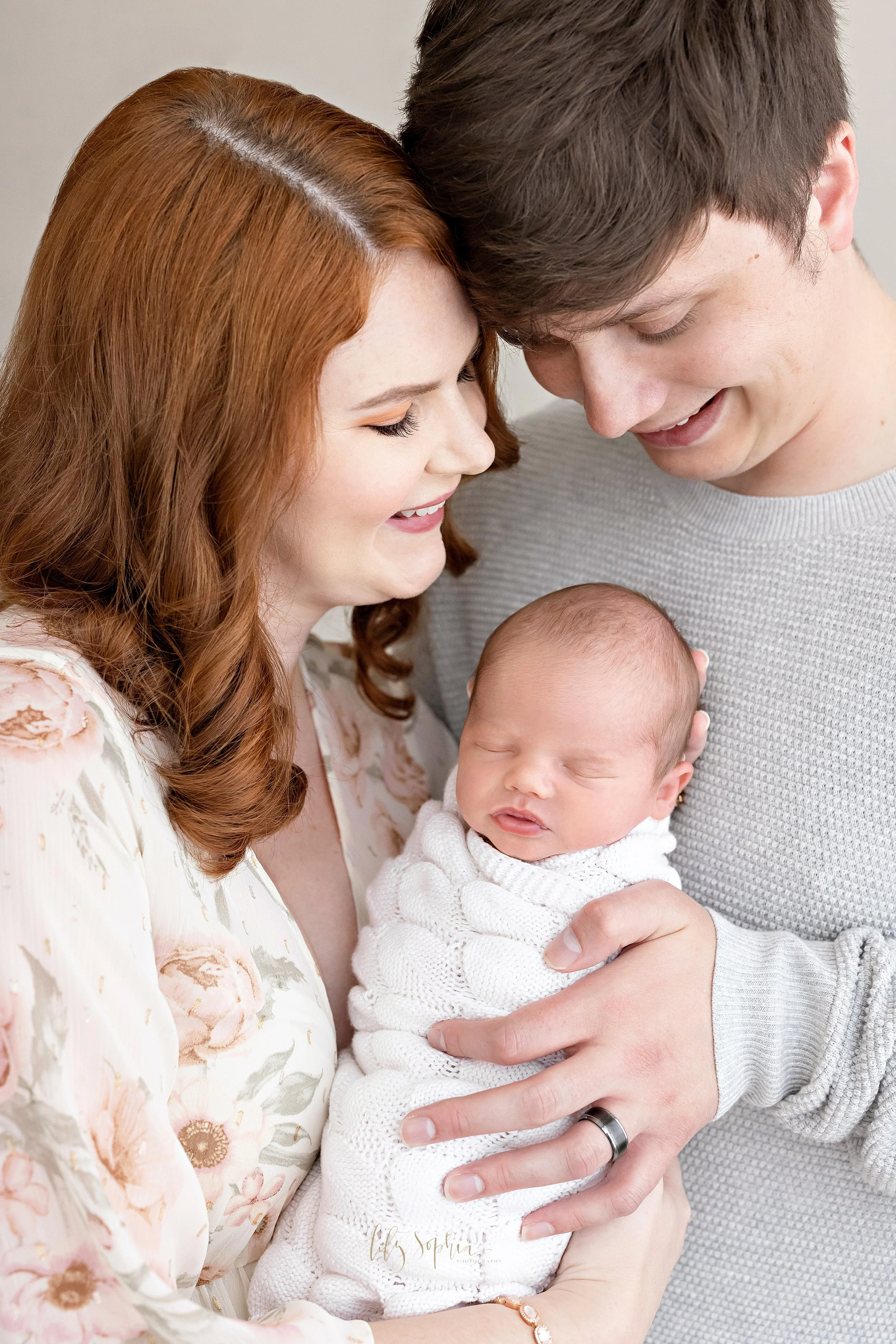  Newborn portrait of an infant boy as he is held by his mother in front of his father’s chest and the two parents marvel at their son taken in a natural light studio near Midtown in Atlanta, Georgia. 