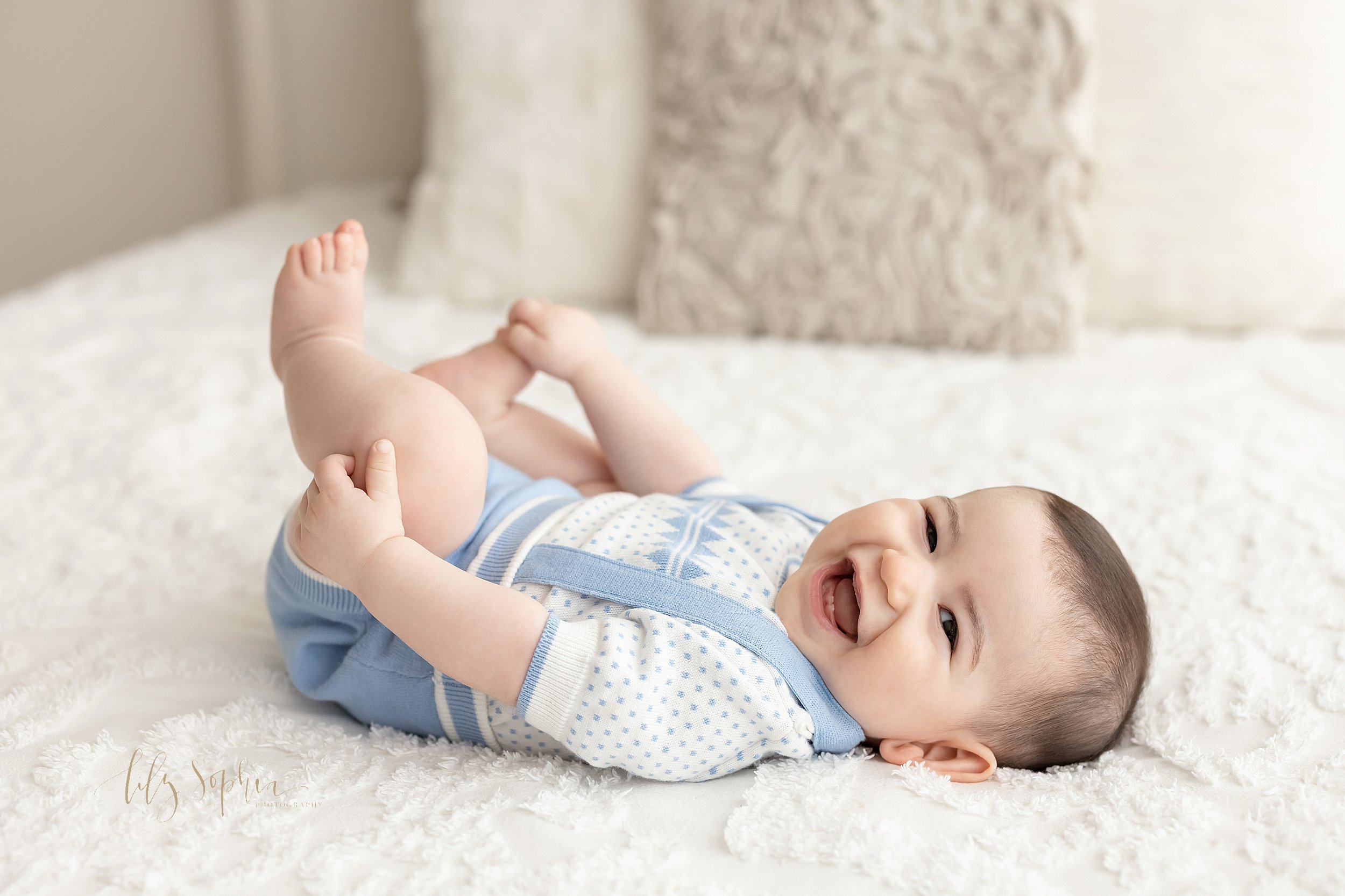  Milestone photo of a six month old little boy as he lies on his back on a bed playing with his feet and laughs taken in a natural light studio near Alpharetta in Atlanta, Georgia. 