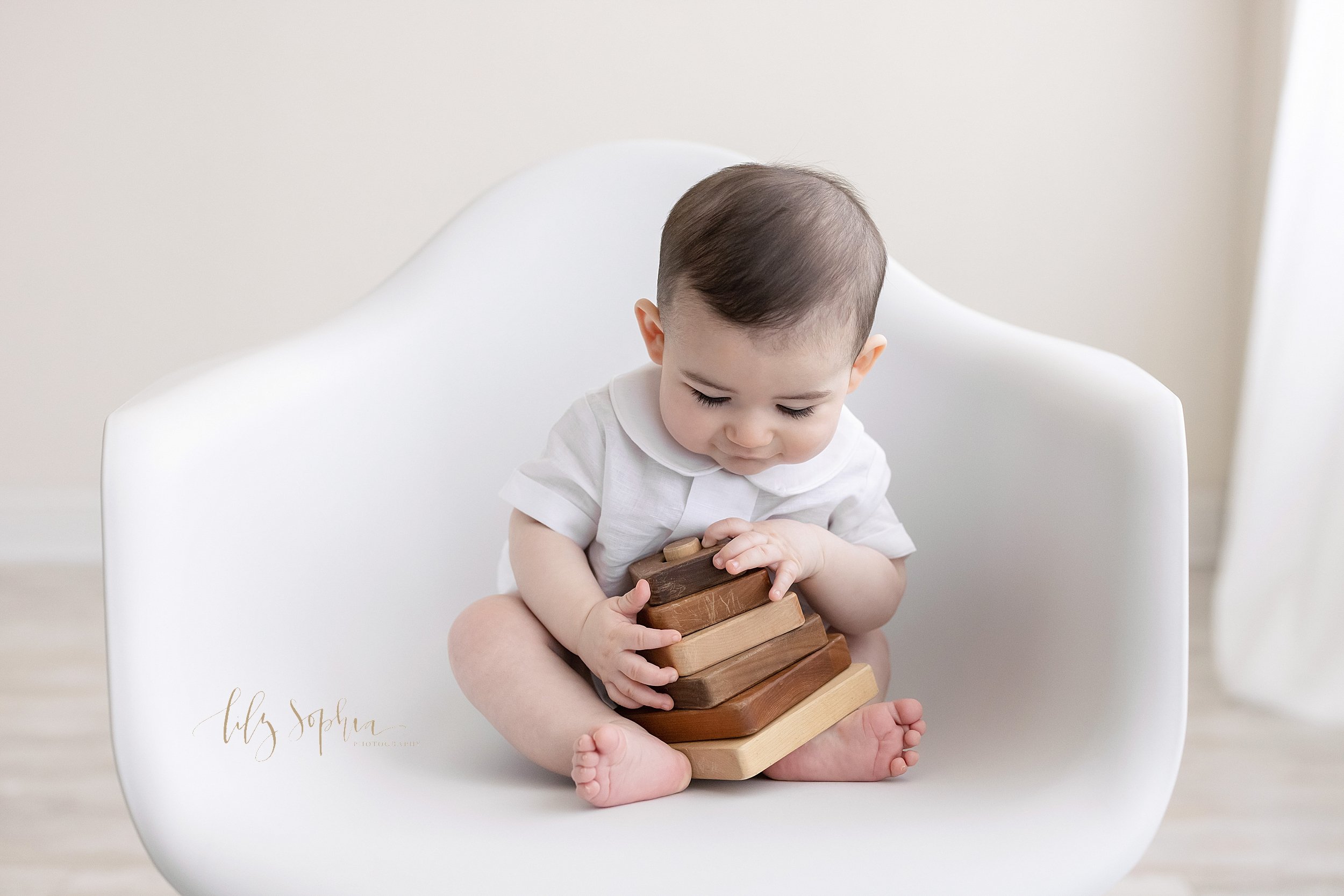  Milestone portrait of a six month old boy as he sits in a white molded chair and plays with a wooden stacking toy taken in front of a window streaming natural light in a studio near Poncey Highlands in Atlanta. 