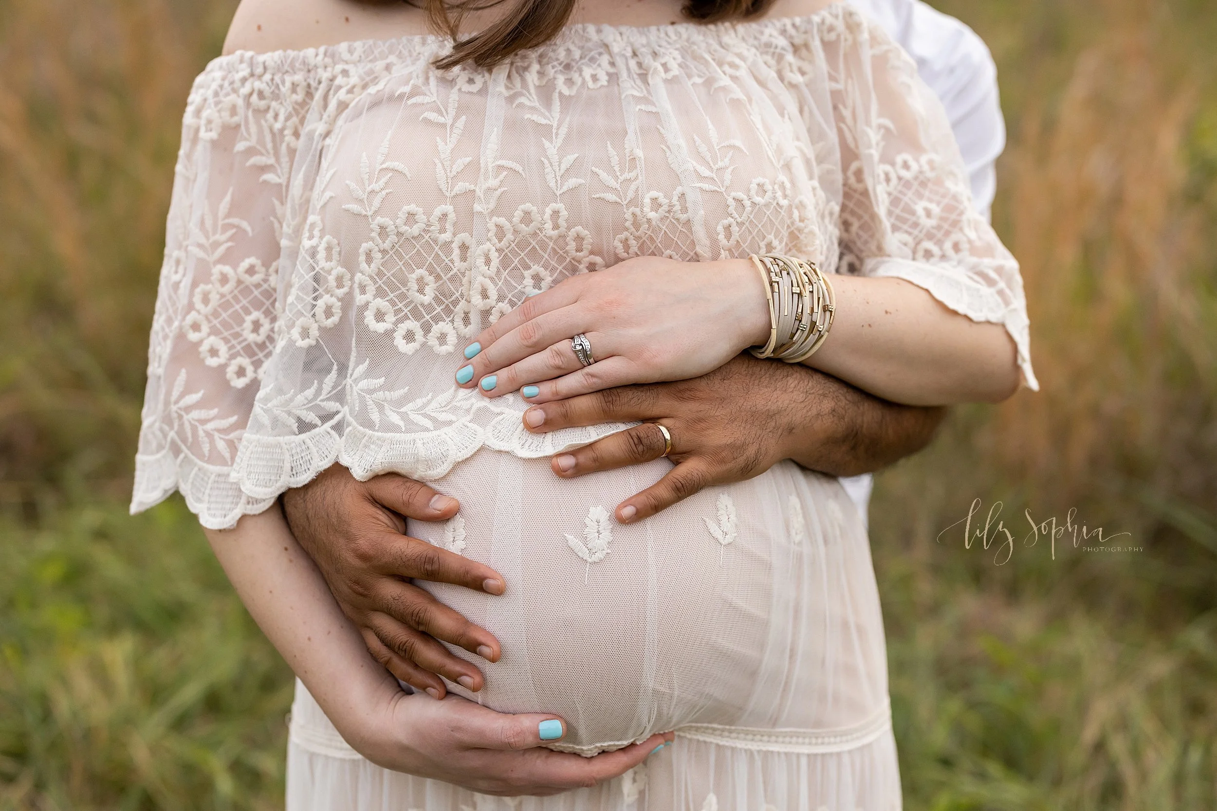  Close-up maternity photo of an expectant mother as she stands with her husband behind her and the two of them place their hands on their child in utero taken in an Atlanta field at sunset. 