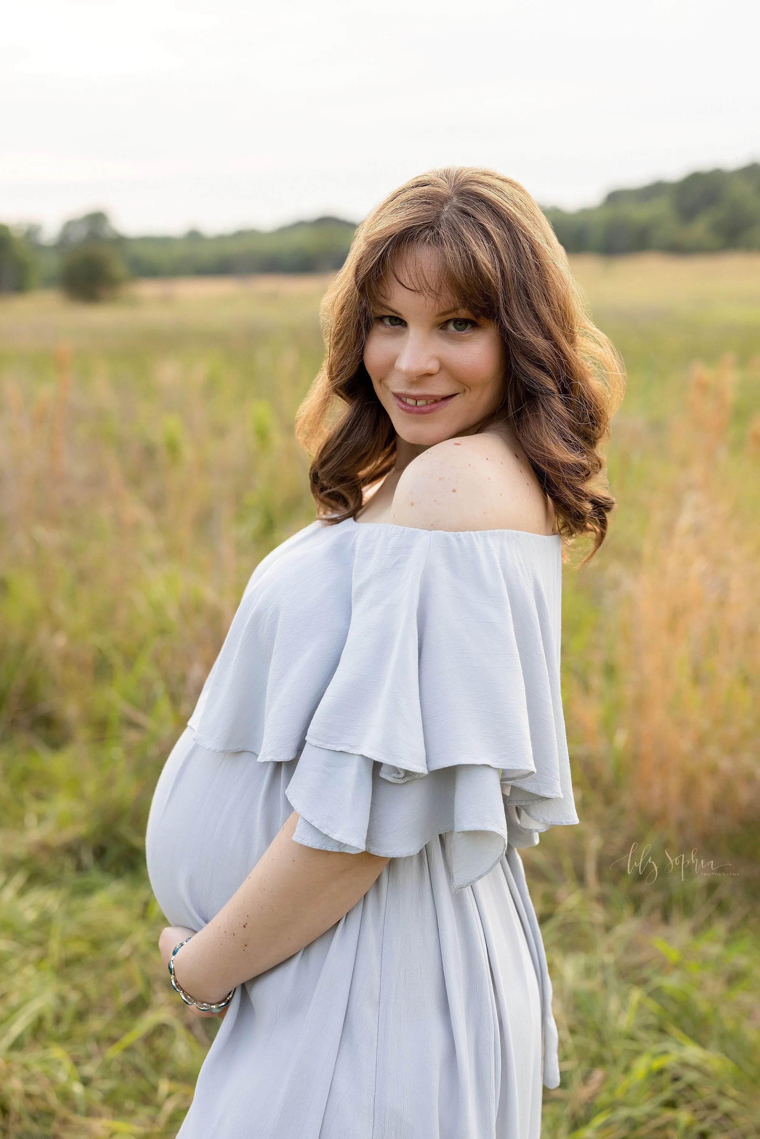  Maternity portrait of a pregnant mother as she stands wearing a full-length gown and looks over her shoulder as she frames her belly taken at sunset in a field near Atlanta. 