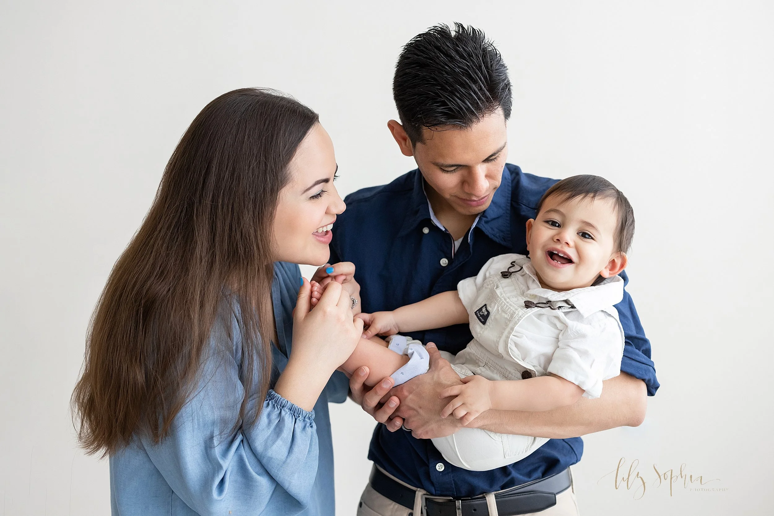  First birthday photos of a smiling one year old boy as he is held in his father’s arms and his mother holds her son’s bare feet while the family stands in a natural light studio near Oakhurst in Atlanta, Georgia. 