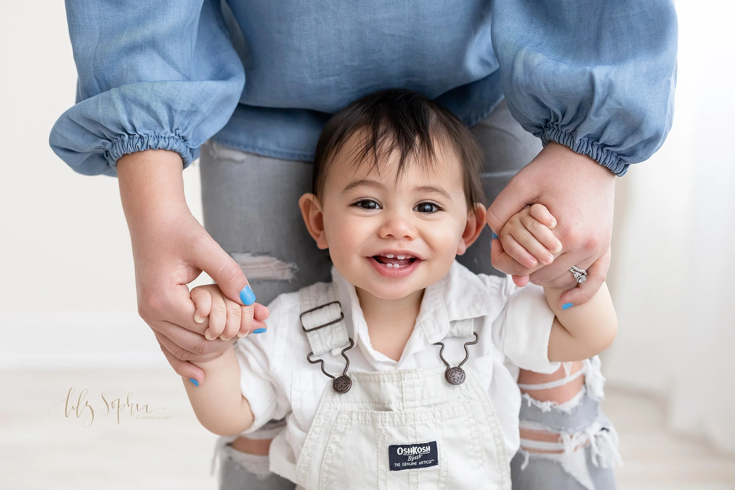 First birthday portrait of a one year old boy showing his teeth as he smiles while dressed in white overalls and a white shirt holding onto his mother’s hands as she walks behind him in a studio using natural light in the Ponce City Market in Atlant