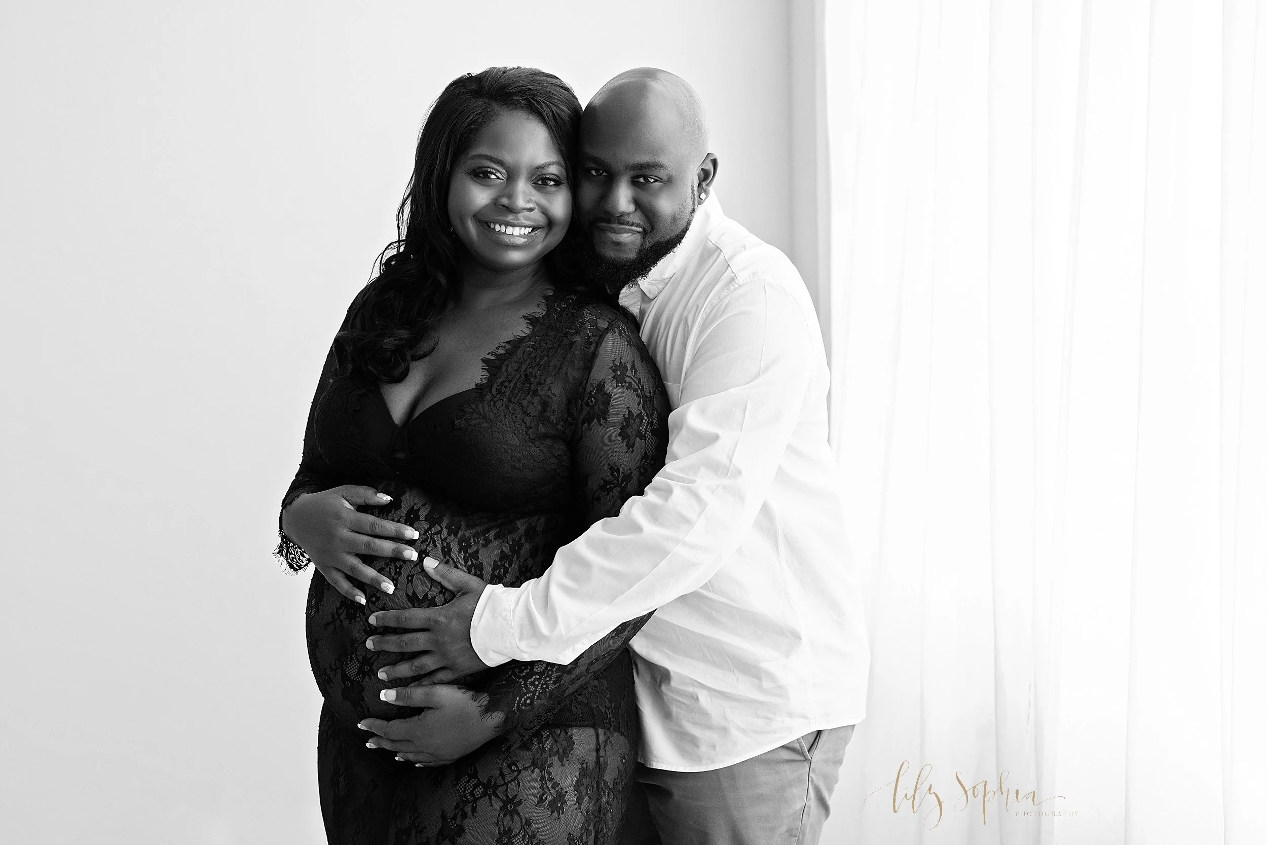  Maternity session of a black couple with the husband standing behind his wife as he places his left hand on his baby in utero and his wife places both of her hands on their baby in utero taken in a studio in Ponce City Market in Atlanta, Georgia. 
