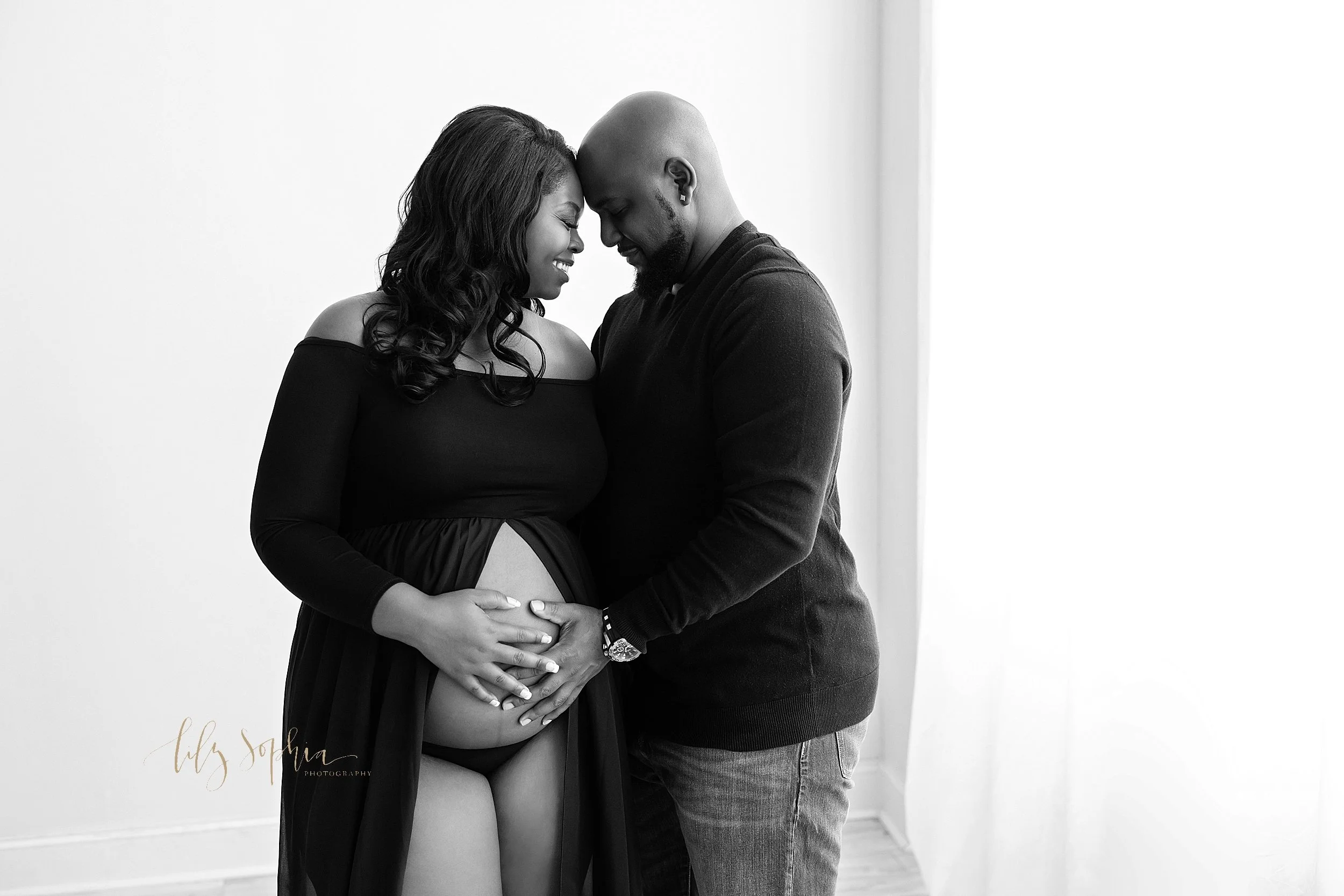  Maternity photo of an African-American couple as the expectant mother wears a black off-the-shoulder, split front knit dress and her husband wears a black long sleeved knit shirt with the couple placing their foreheads together and each of them plac