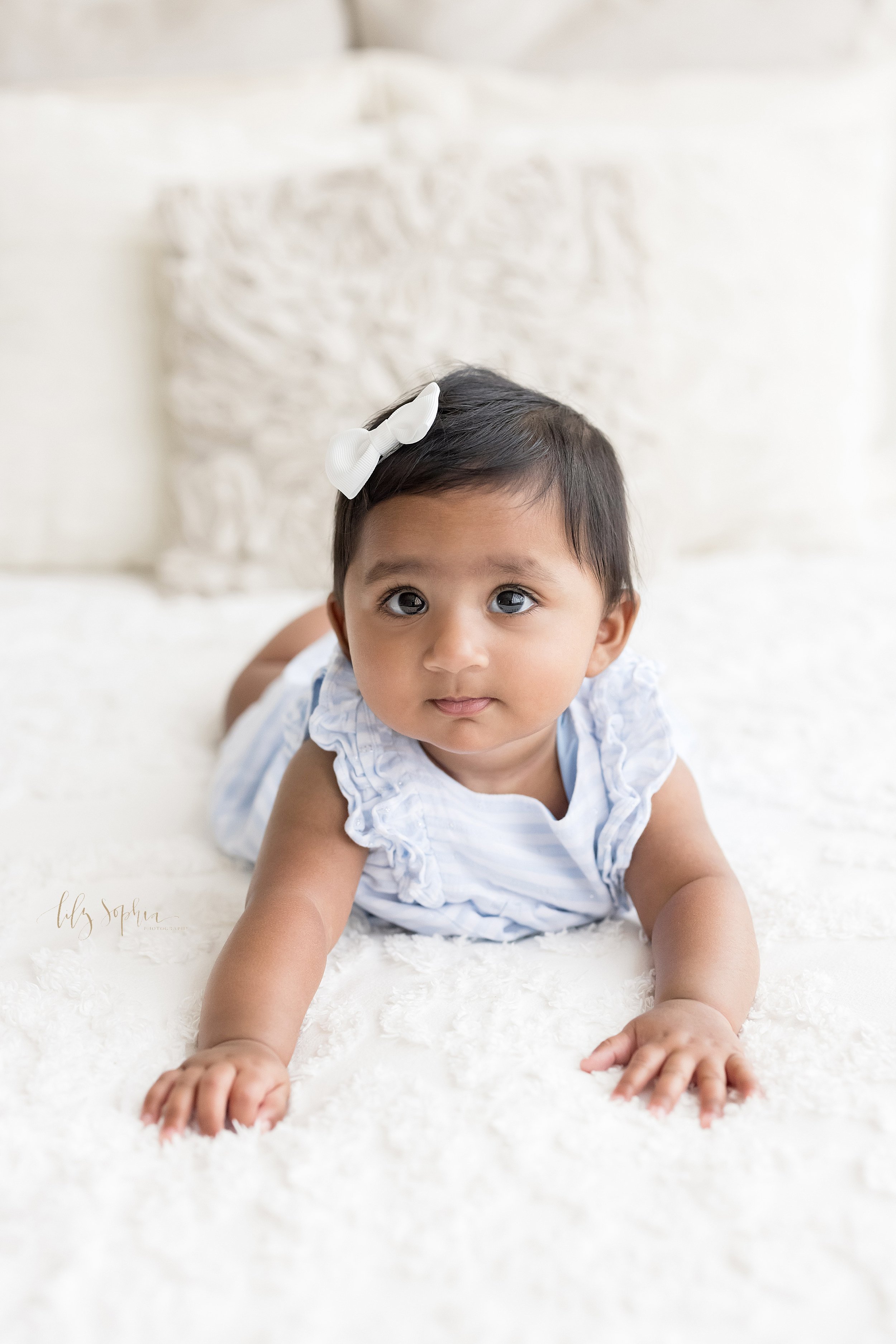  Portrait of a six month old Indian girl as she lies on her stomach on a bed with a white bow in her dark hair in a studio near Buckhead in Atlanta that uses natural light. 