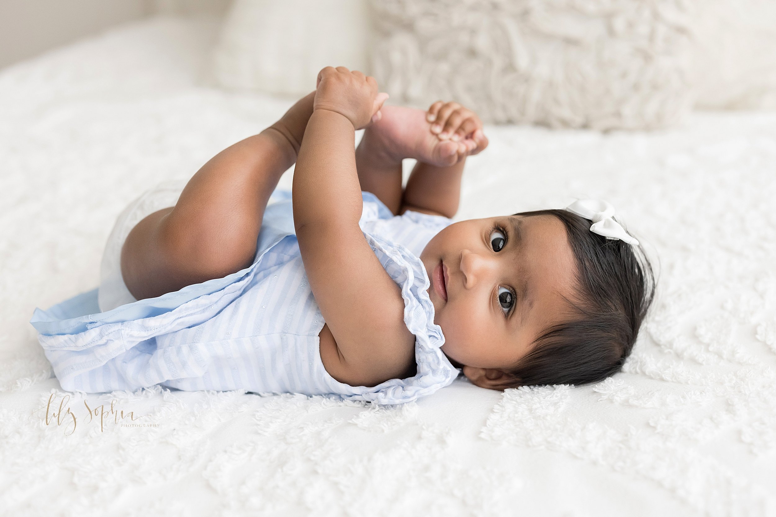  Milestone portrait of a six month old Indian girl as she lies on her back on a bed and plays with her feet taken in a natural light studio in the Ponce City Market in Atlanta, Georgia. 