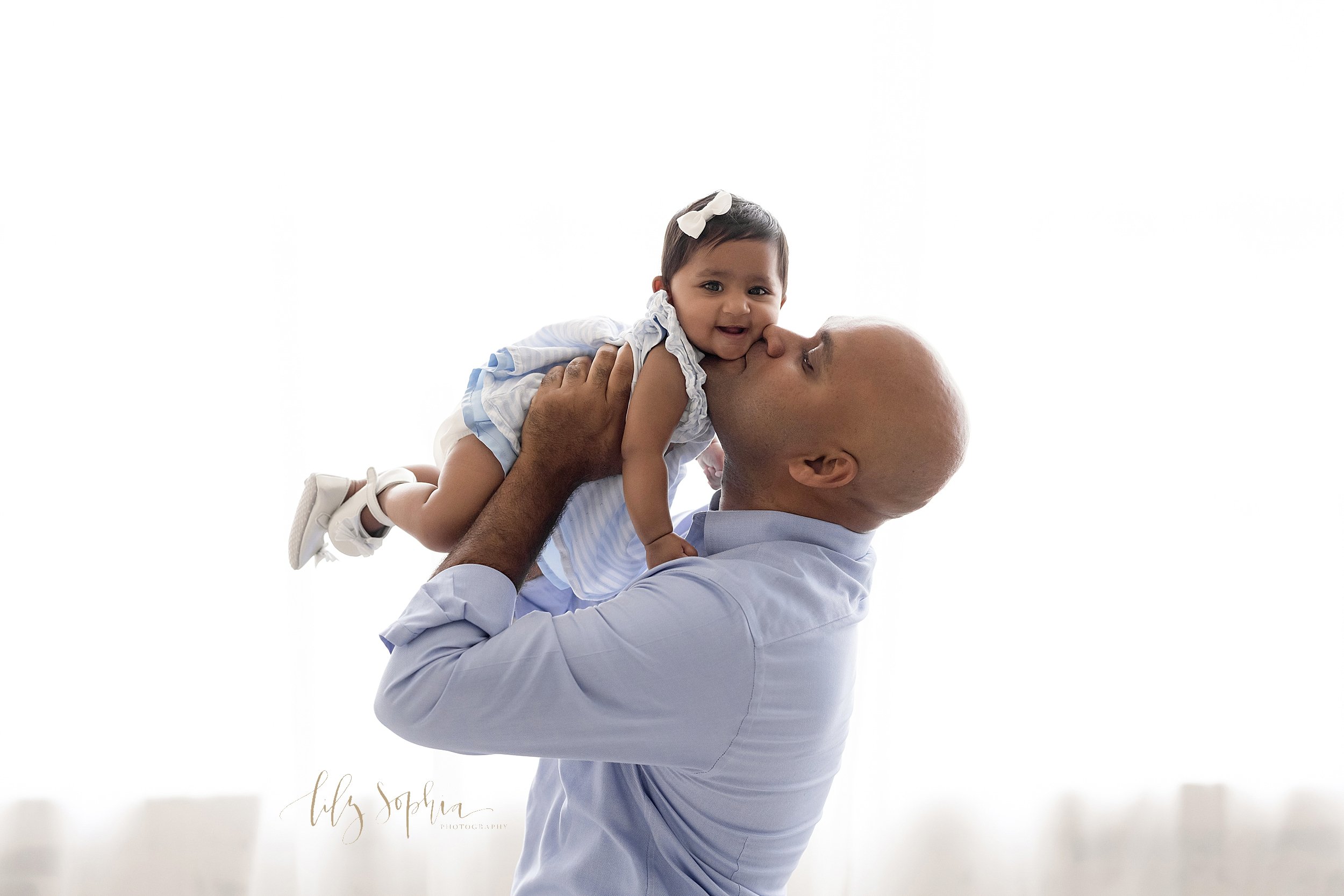  Family photo of an Indian father as he holds his six month old daughter above his head and kisses her on the cheek while standing in front of window streaming natural light in a studio near Kirkwood in Atlanta. 