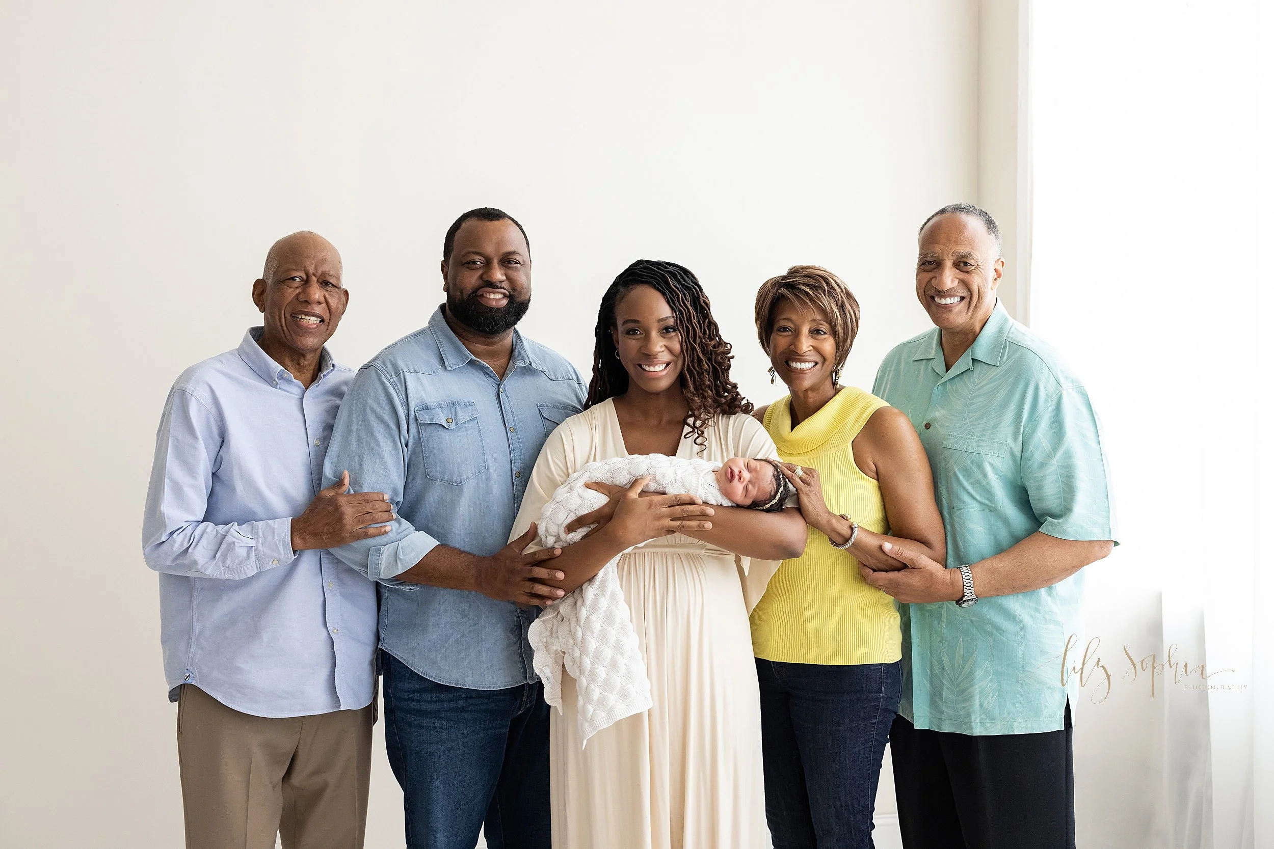  Intergenerational family portrait of a family with mom cradling her newborn in her arms, dad standing to mom’s left and placing his hand on mom’s elbow, and dad’s dad to the left of the newborn’s father with his hand on dad’s elbow, mom’s mom to the