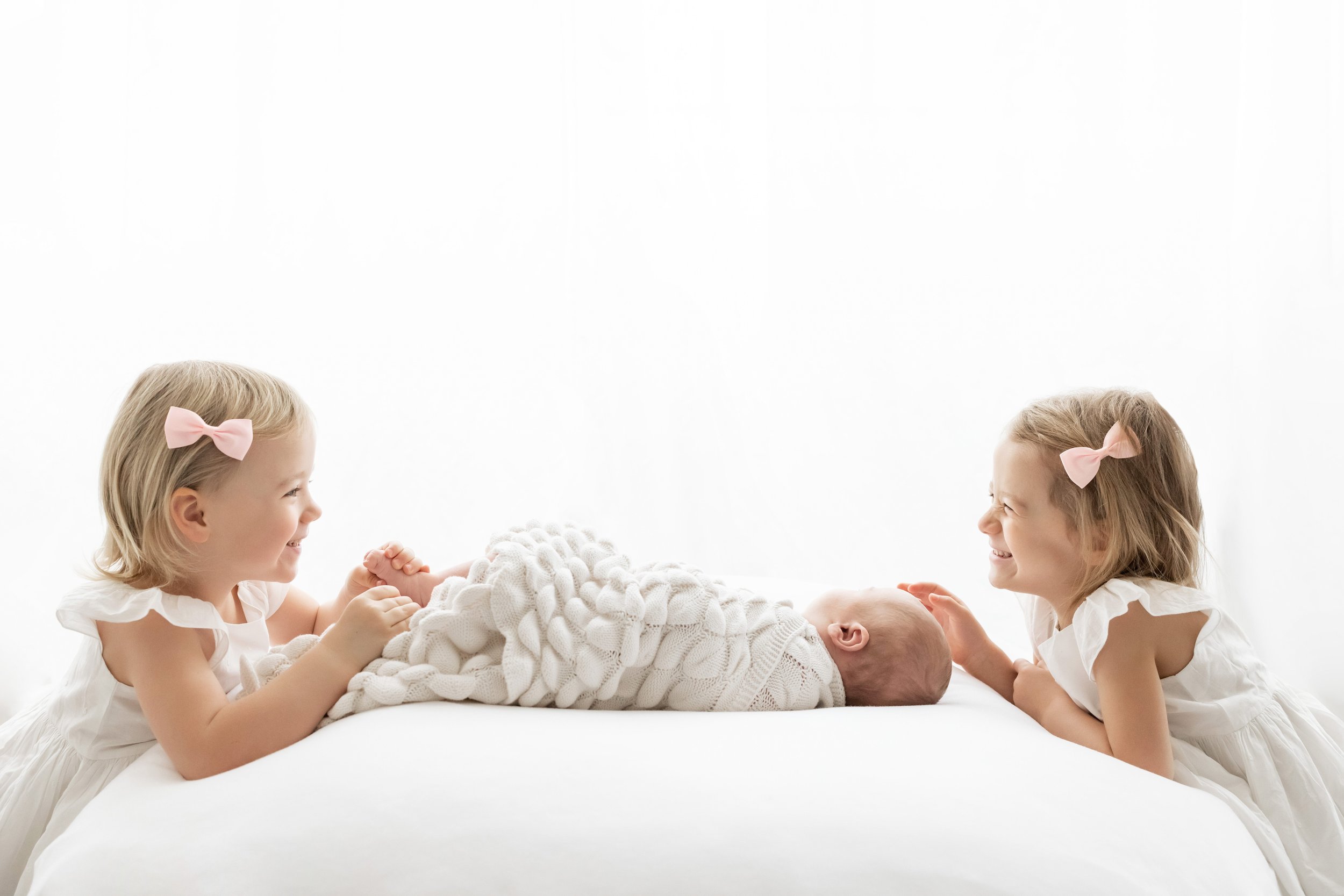  Family newborn portrait of an infant lying on his back swaddled to his chin in a soft white blanket and his two sisters kneel next to the bed, one at his head and the other at his feet taken in an Atlanta studio utilizing natural light. 