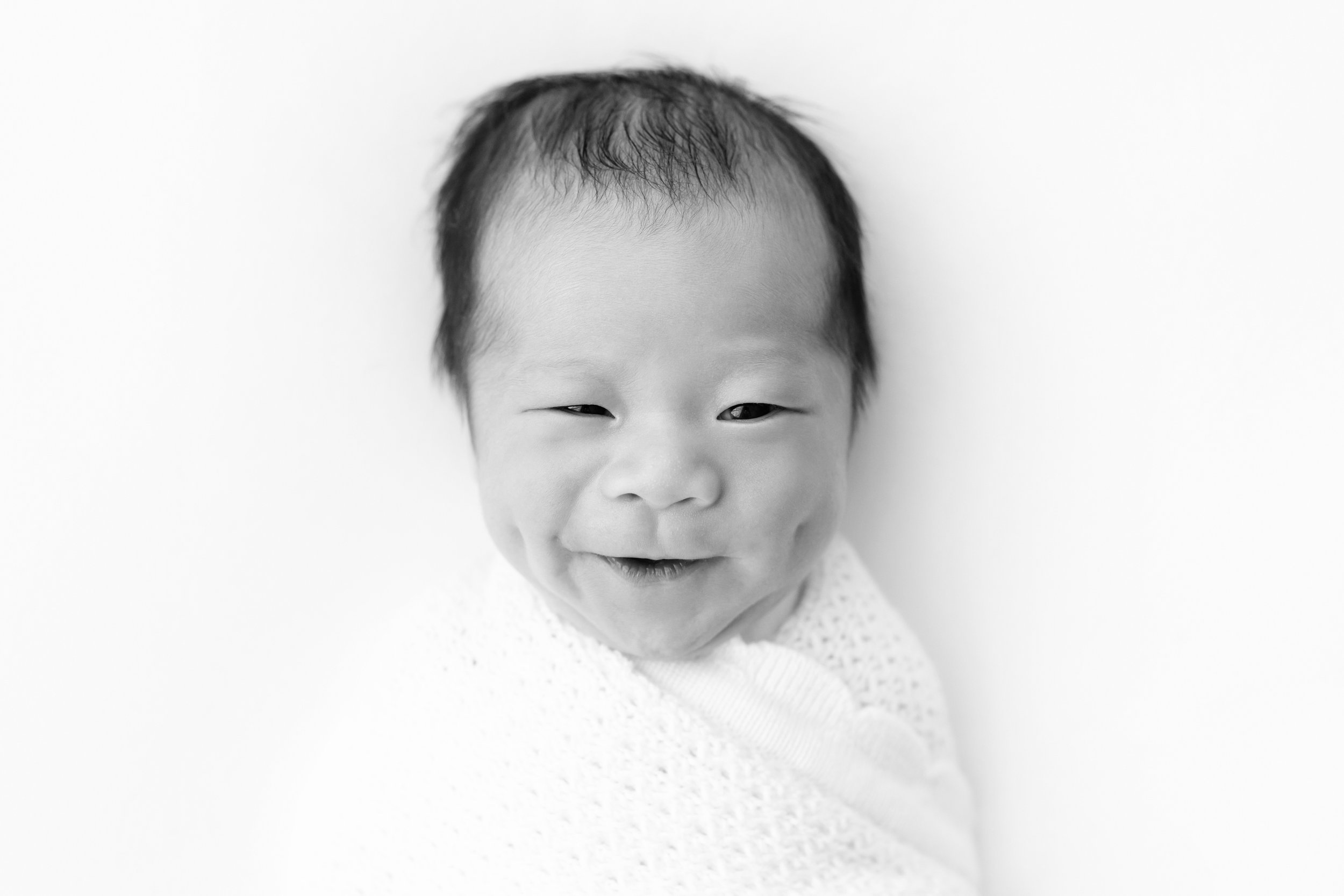  Newborn Asian baby smiles highlighting his dimples while awake and swaddled to his chin in a soft white blanket in a natural light studio near Alpharetta in Atlanta. 