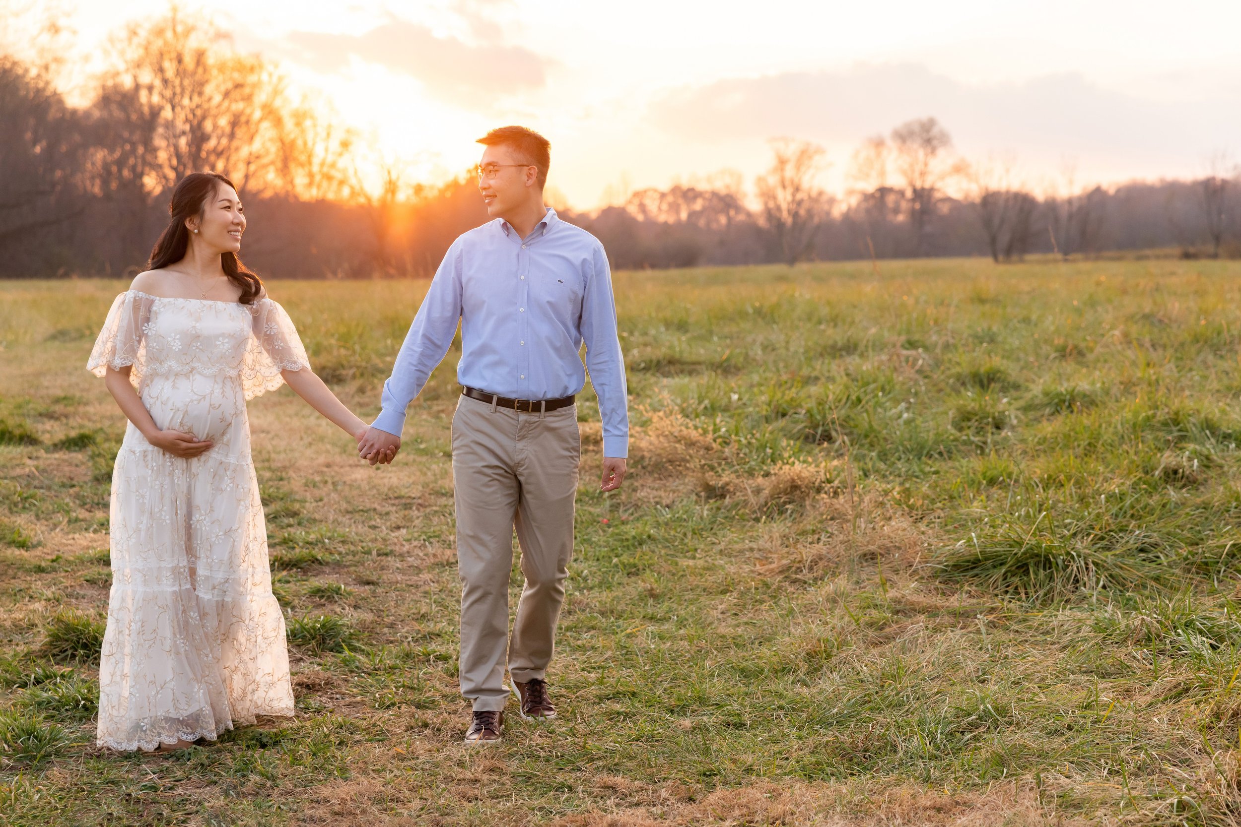  An expectant Asian couple walk hand-in-hand at sunset in a field near Atlanta with the mother holding her baby in utero with her right hand as she wears a flowing full-length gown and her husband wears khaki’s and a long-sleeved blue shirt. 