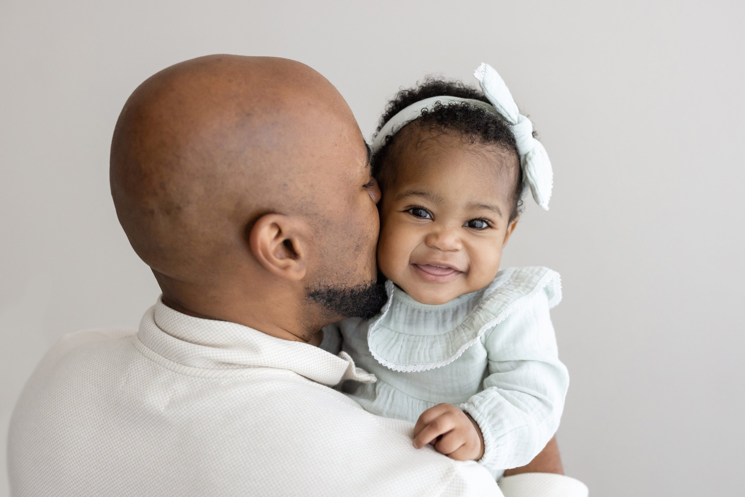  An African-American father holds his young daughter in his arms and kisses her on the cheek in a studio near Morningside in Atlanta as she peeks over his shoulder and giggles. 