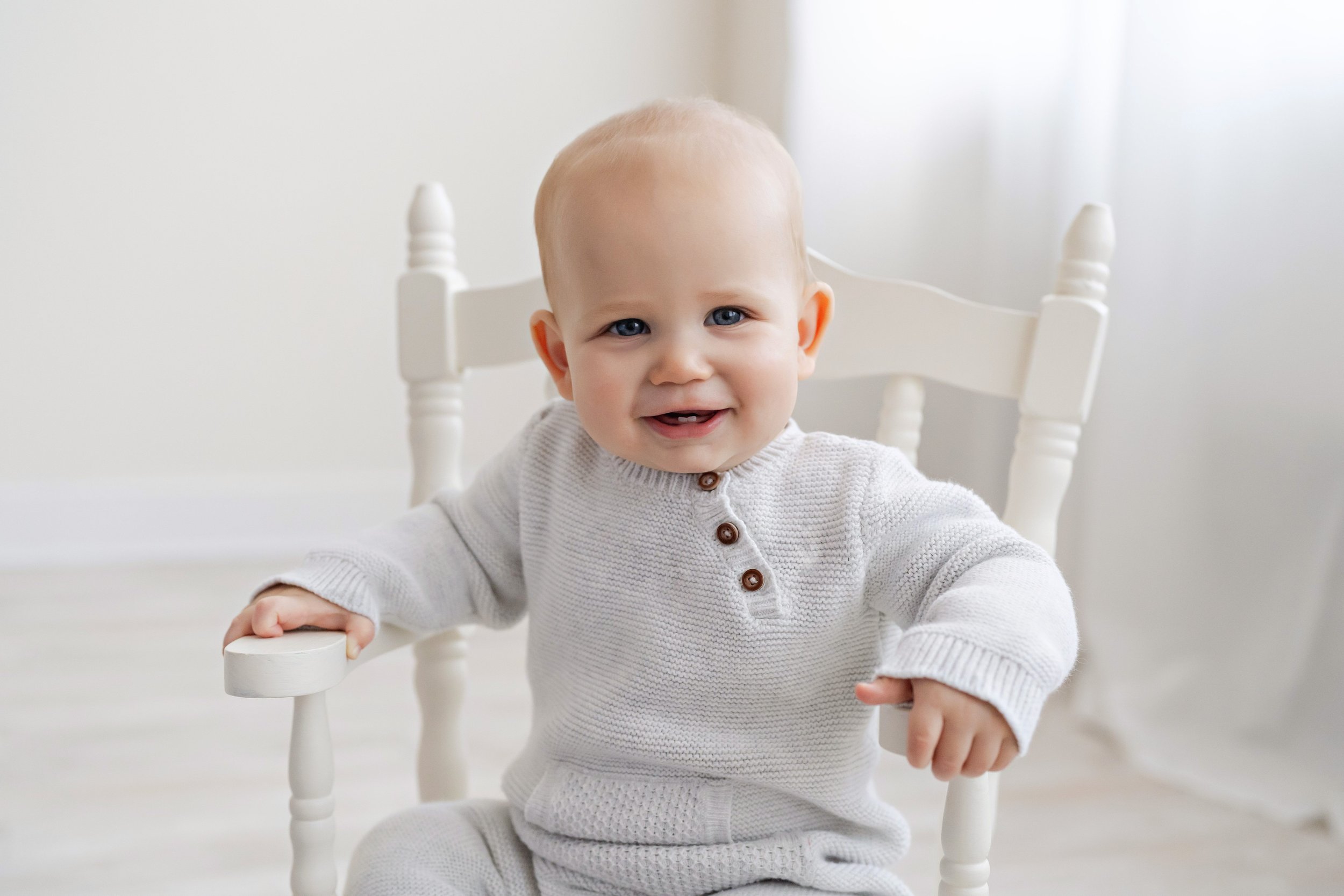  A proud one year old sits in a wooden rocking chair in Lily Sophia Photography Studios in Atlanta and shows his front teeth.  