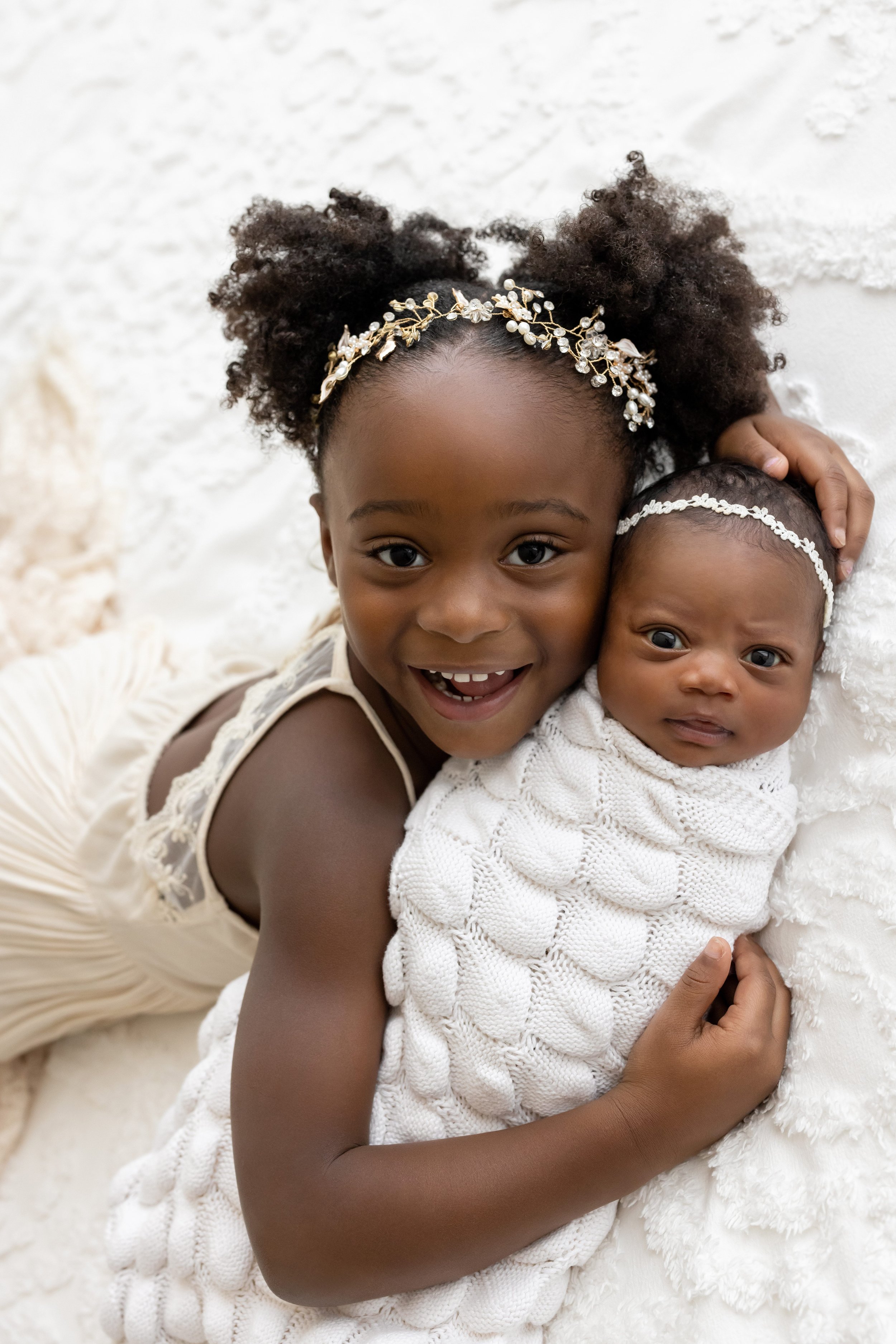  Newborn sibling photo with a sister holding her wide awake newborn sister who is swaddled to her chin with a soft white blanket and wearing a delicate headband on her head as they lie together on a bed in a studio near Midtown in Atlanta in natural 