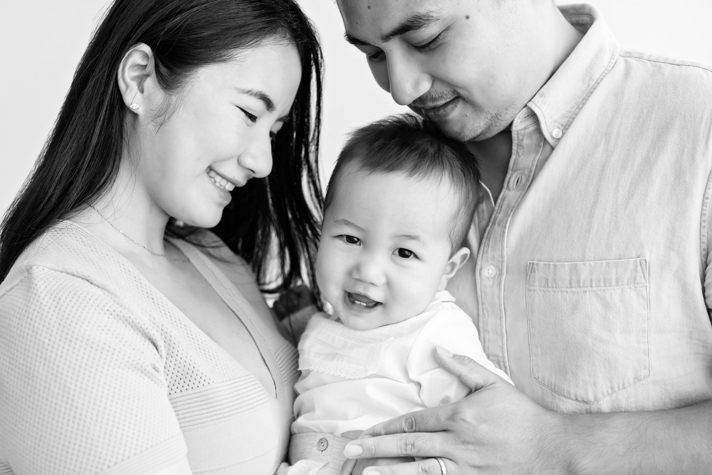  An Asian mother and father hold their son between them while he smiles and shows his tiny front teeth in a studio near Midtown in Atlanta, Georgia. 