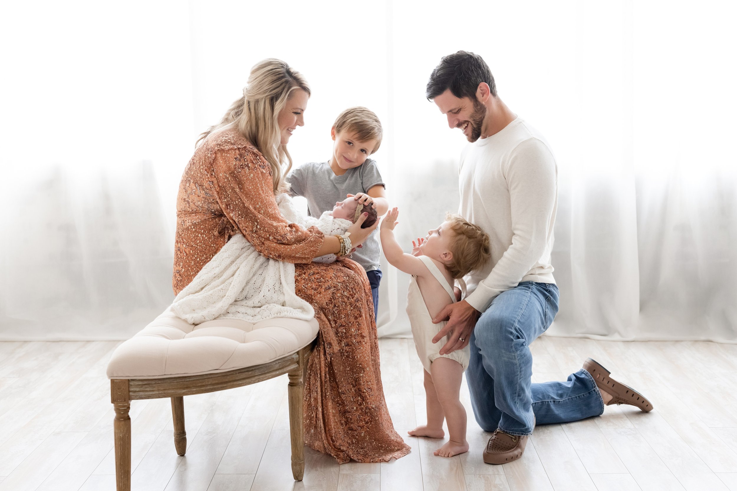  Family photography session of a newborn with mom holding the infant on her lap, her son standing to her side looking on, her husband kneeling on one knee facing her and steadying their toddler as the toddler touches the head of their newborn taken i