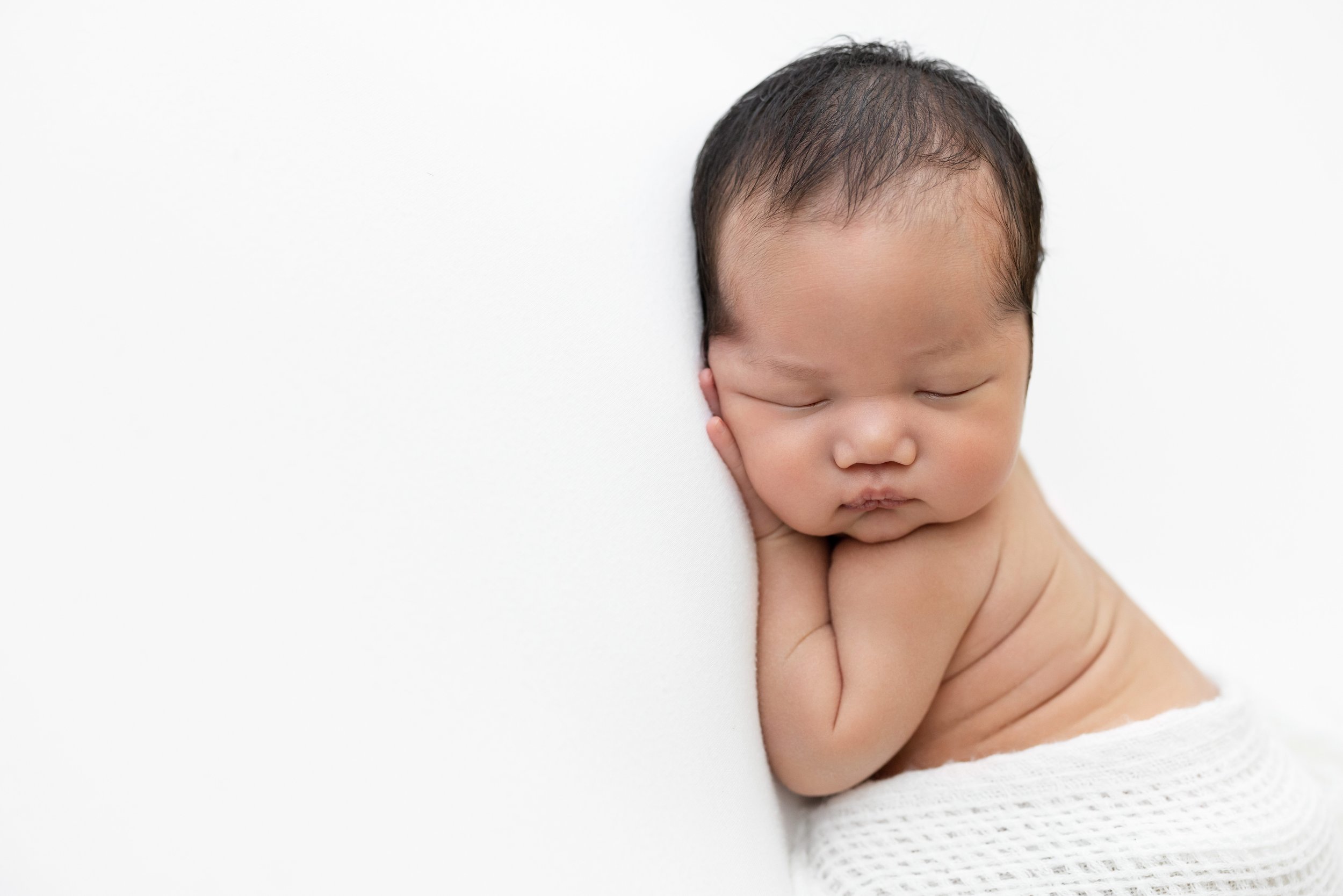  An Asian newborn lies on his stomach with his right hand under his chubby cheek and the folds in his back can be seen as he peacefully sleeps in a natural light studio near Old Fourth Ward in Atlanta. 