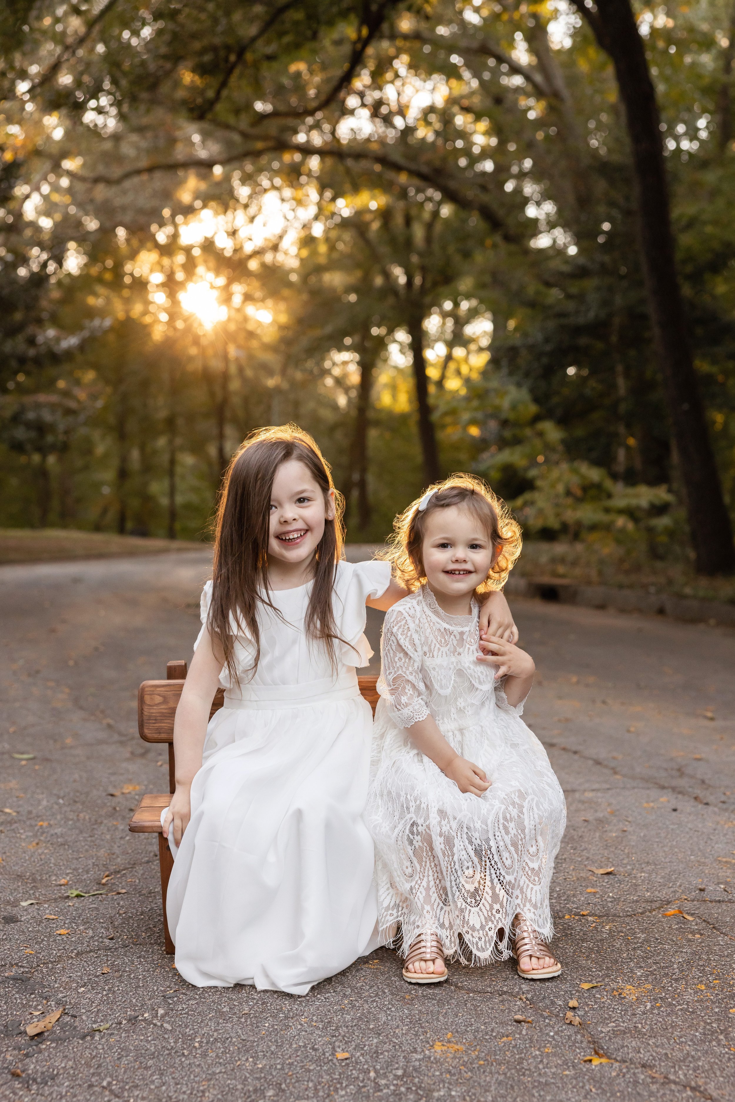  Family portrait of two sisters as they sit on a bench along a path in an Atlanta park as the older sister puts her and around the younger sister’s shoulder and the younger sister holds her hand with the sun setting behind them. 