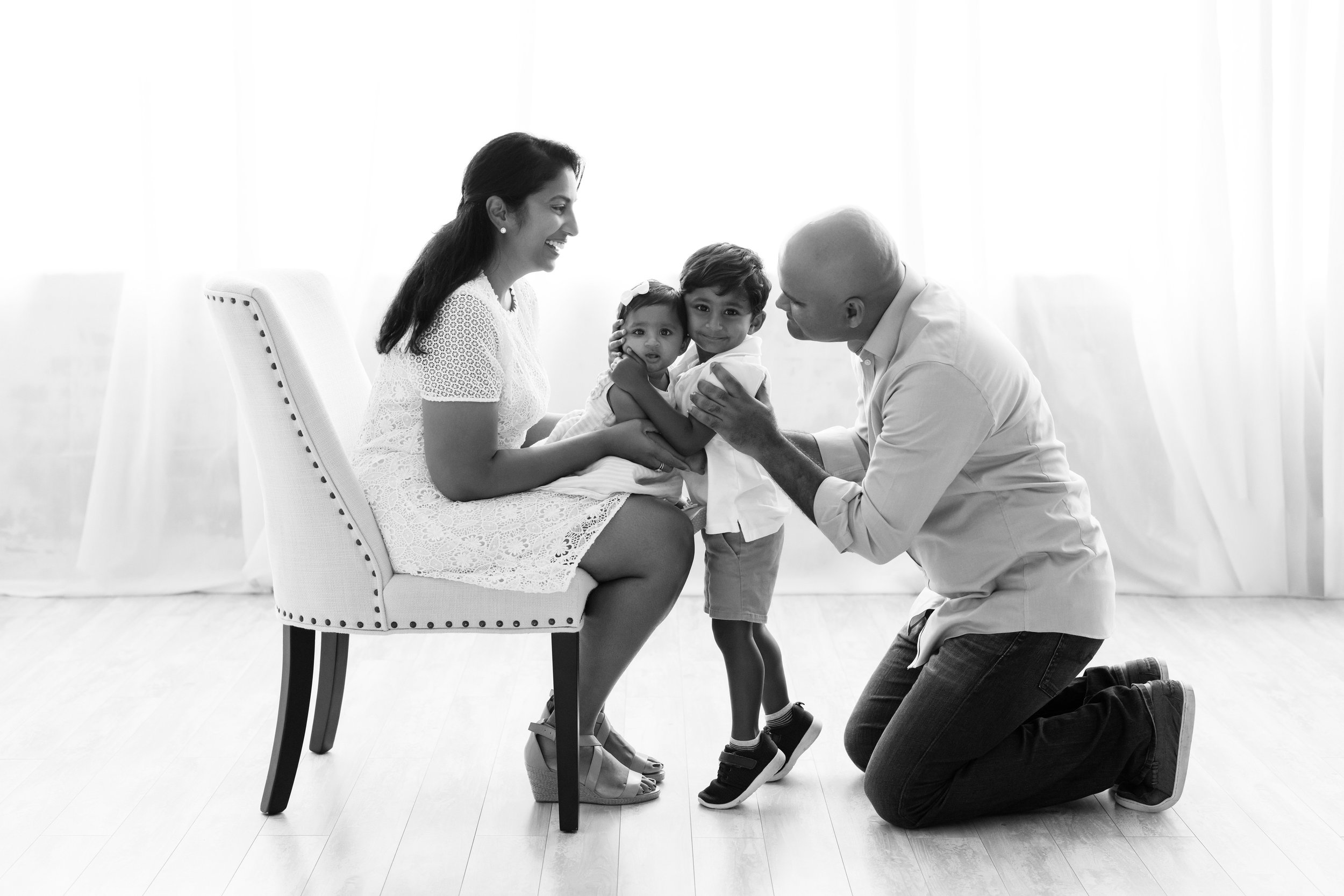  An Indian mother holds her young daughter on her lap as she sits in a chair in front of a natural light window and her young son hugs his sister as dad kneels behind the son to steady him taken in a studio near Vinings in Atlanta. 