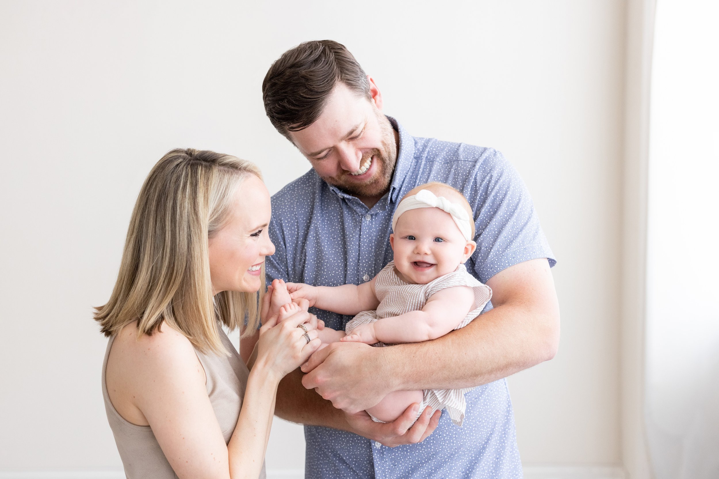  A father holds his young daughter in front of him as mom tickles her bare feet while the family stands in front of a window streaming natural light in a studio near Kirkwood in Atlanta, Georgia. 