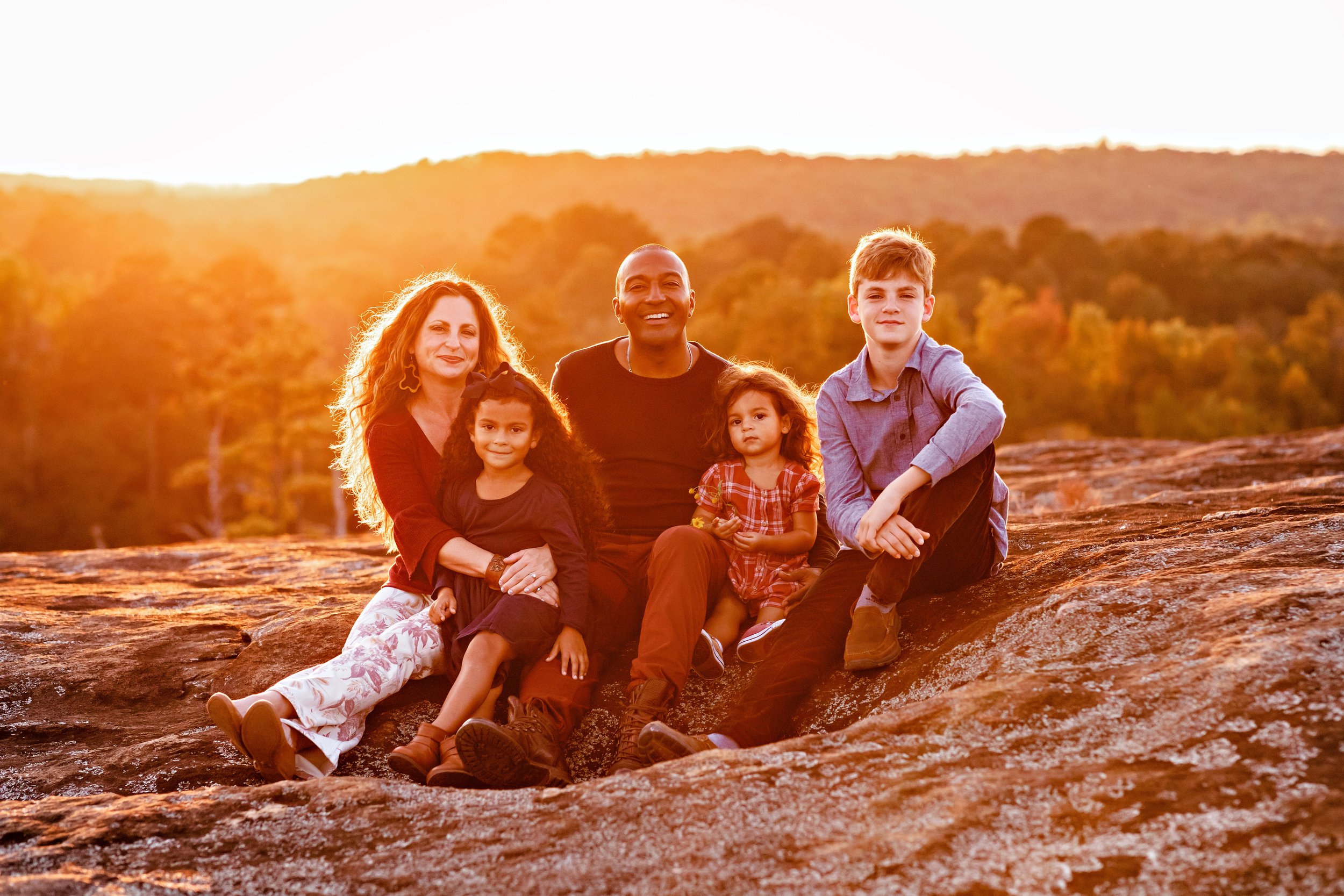  Family portrait of a mother with her hands wrapped around her daughter, a father sitting next to his wife with their younger daughter on his lap and their older son sitting next to their dad on a mountaintop at sunset near Atlanta. 