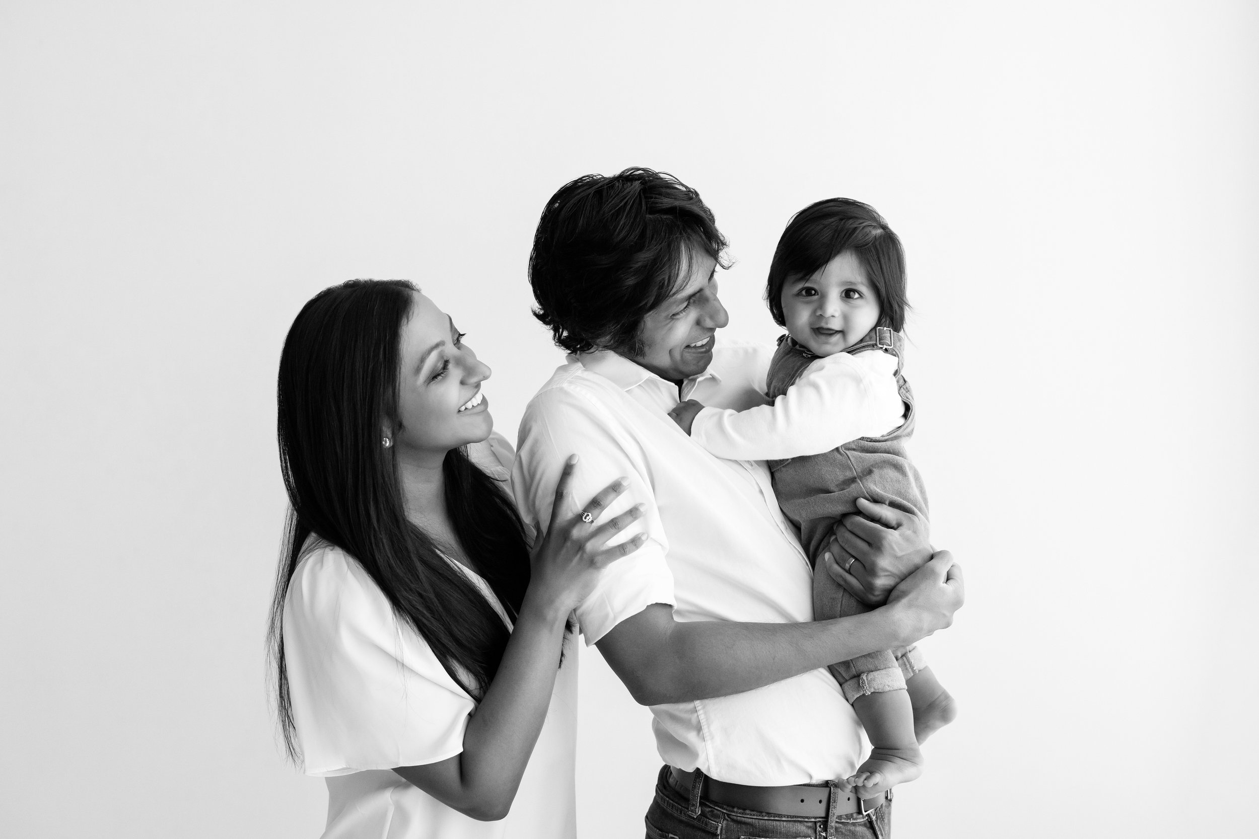  Family portrait of an Indian family with dad holding their year old son and mom standing behind dad looking on as their son smiles taken in a natural light studio near Alpharetta in Atlanta. 