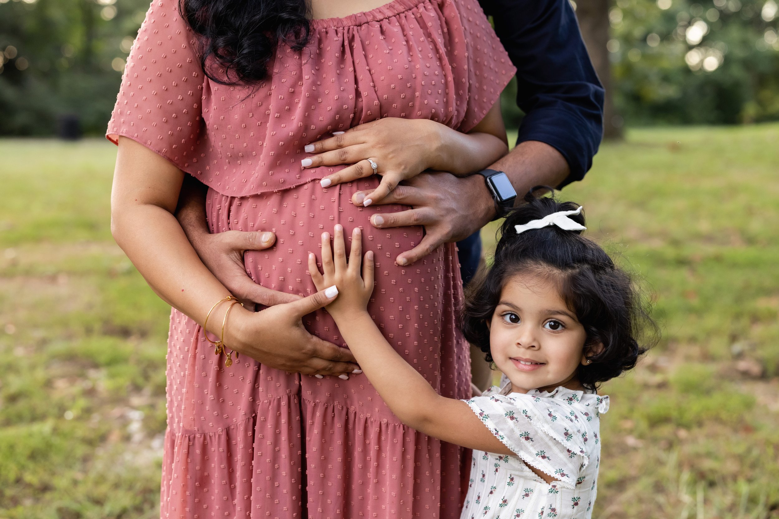  At a park near Atlanta a father stands behind his expectant wife and the father and mother place their hands on their child in utero as their young daughter stands next to them and places her hand on her mom’s belly at sunset. 