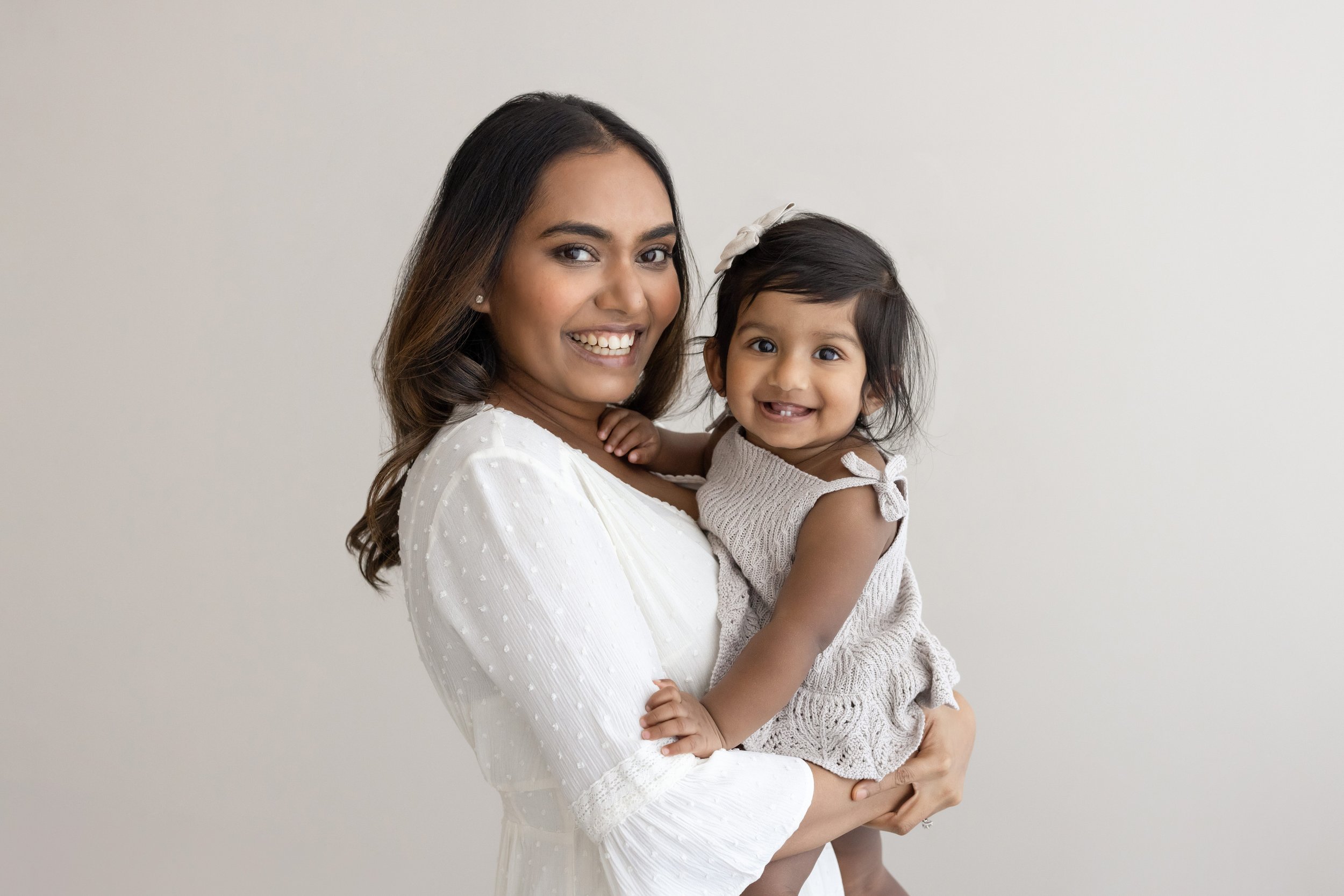  A mother stands wearing a white shirt with belled sleeves holding her one year old daughter, wearing a sundress and a bow in her hair, in her arms as the two of them smile in a natural light studio near Decatur in Atlanta, Georgia. 