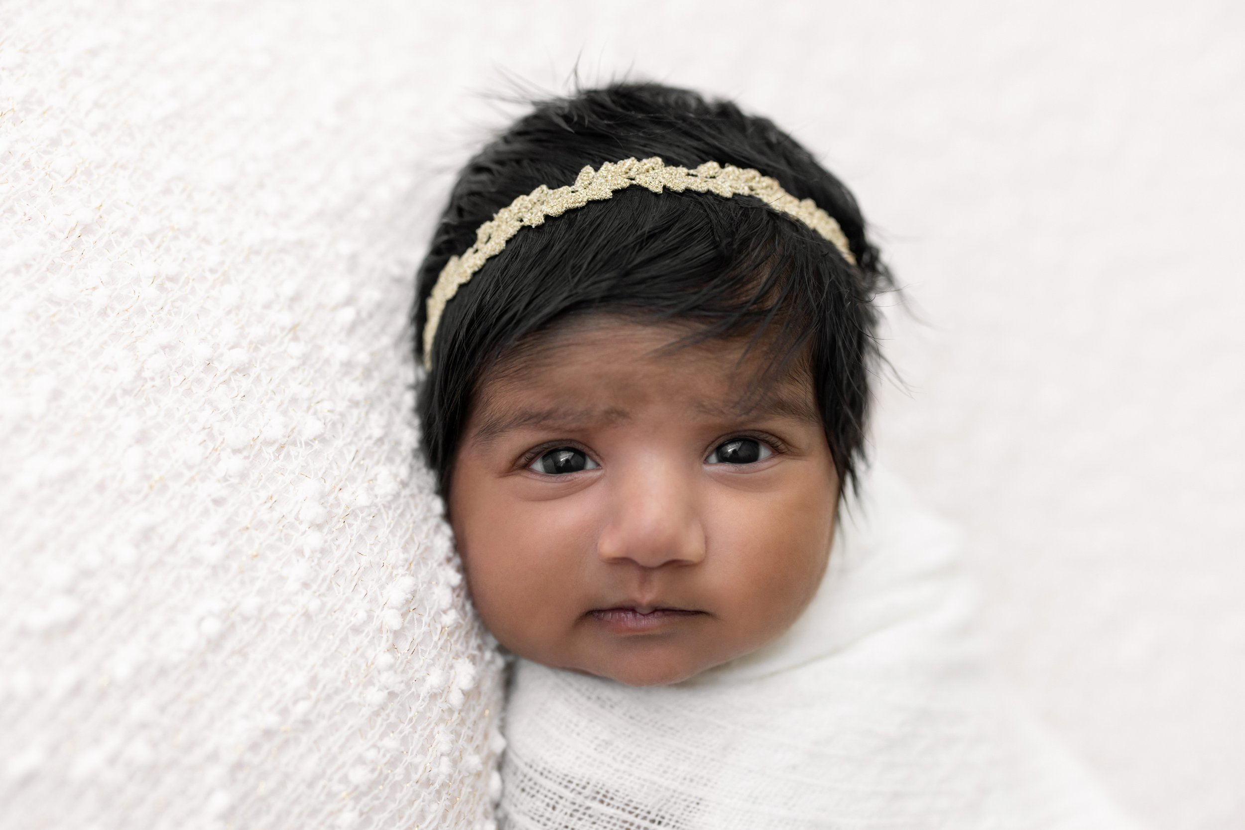  Newborn portrait of a wide awake Indian infant girl as she lies on her back swaddled to her chin with a soft white blanket and wears a delicate headband in her hair taken in a natural light studio near Old Fourth Ward in Atlanta. 