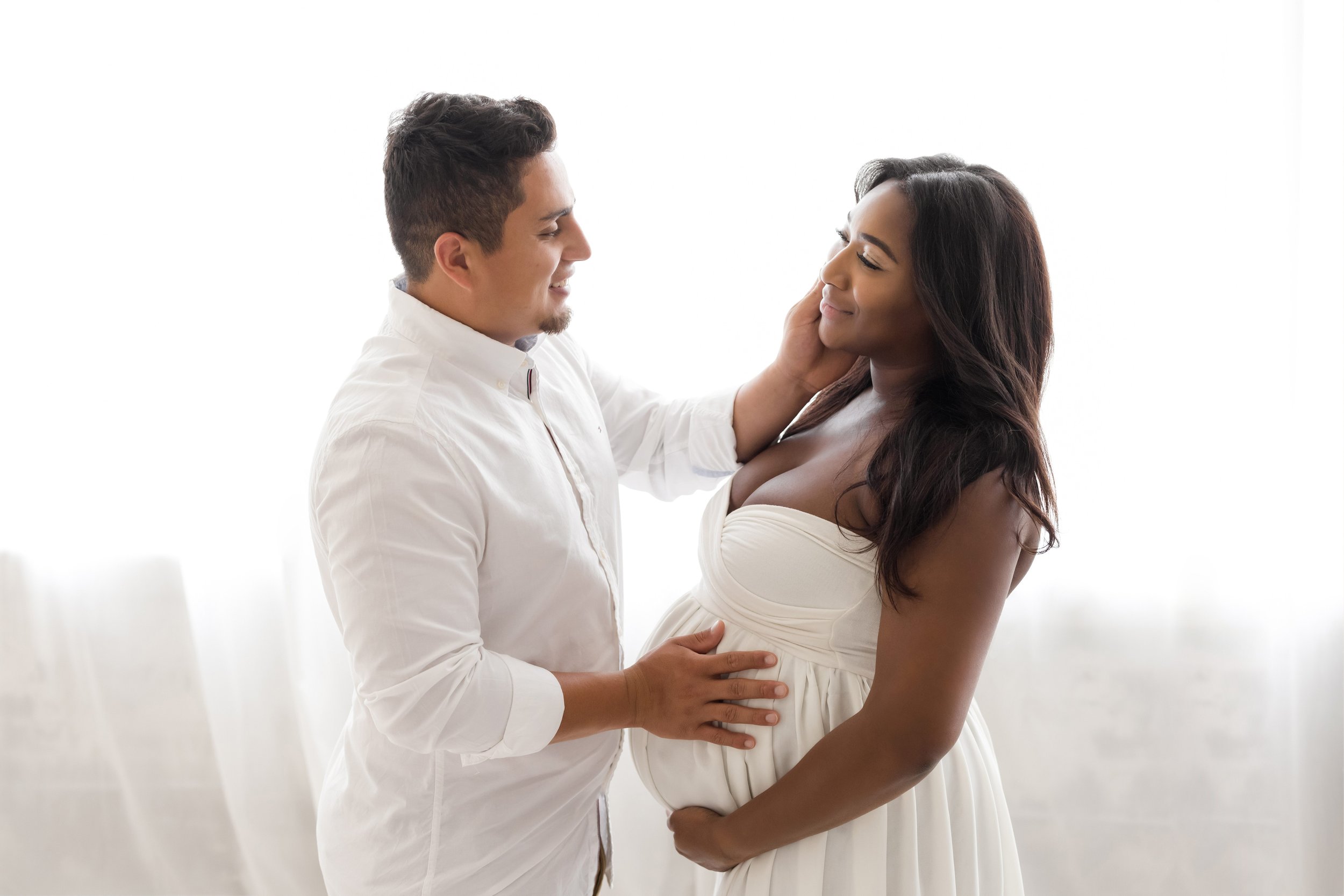  A husband stands facing his wife and places his left hand on his pregnant wife’s cheek while he places his right hand on her belly and the two contemplate the pending birth of their child.  The wife is wearing a strapless white maternity gown as she