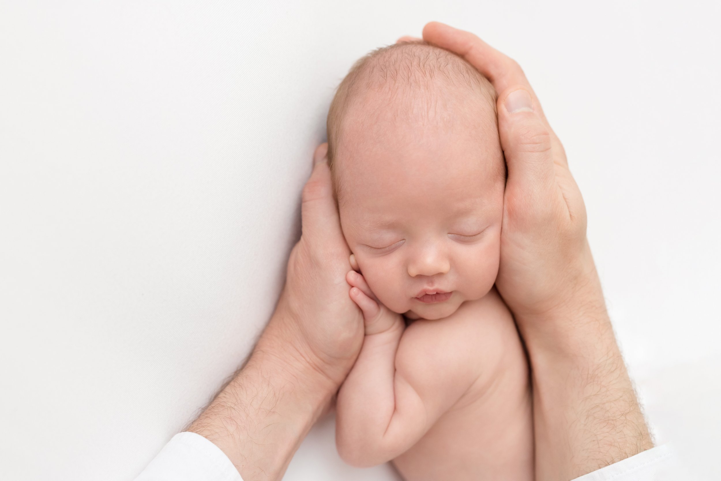  Portrait of a newborn lying on his stomach peacefully sleeping as his father places his son’s head in his hands taken near Brookhaven in Atlanta in a natural light photography studio. 