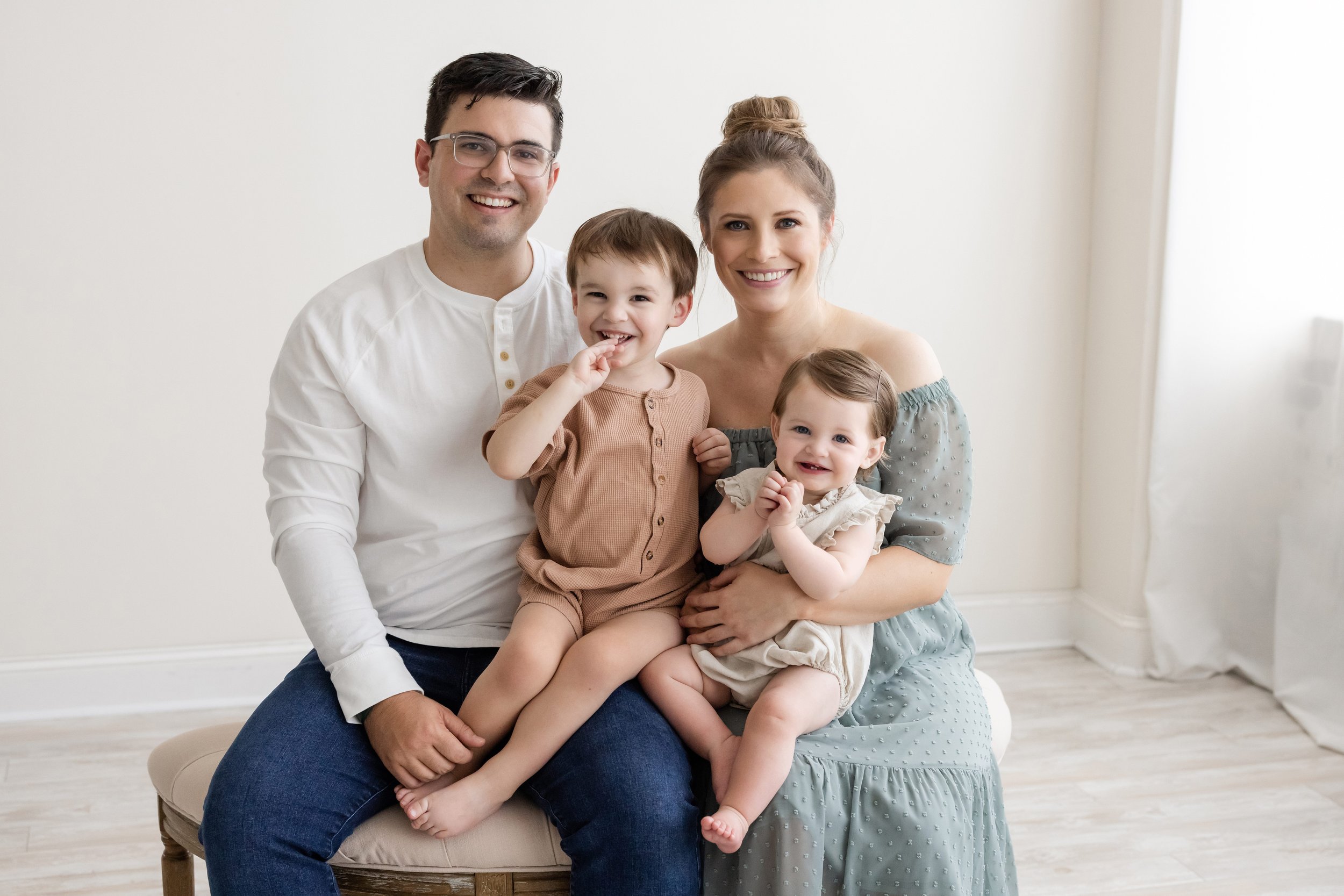  A father and mother sit side by side on a tufted bench in a photography studio with their young son sitting on his father’s left knee and their toddler daughter sitting on mom’s lap.  The family portrait was taken by Lily Sophia Photography near Bro