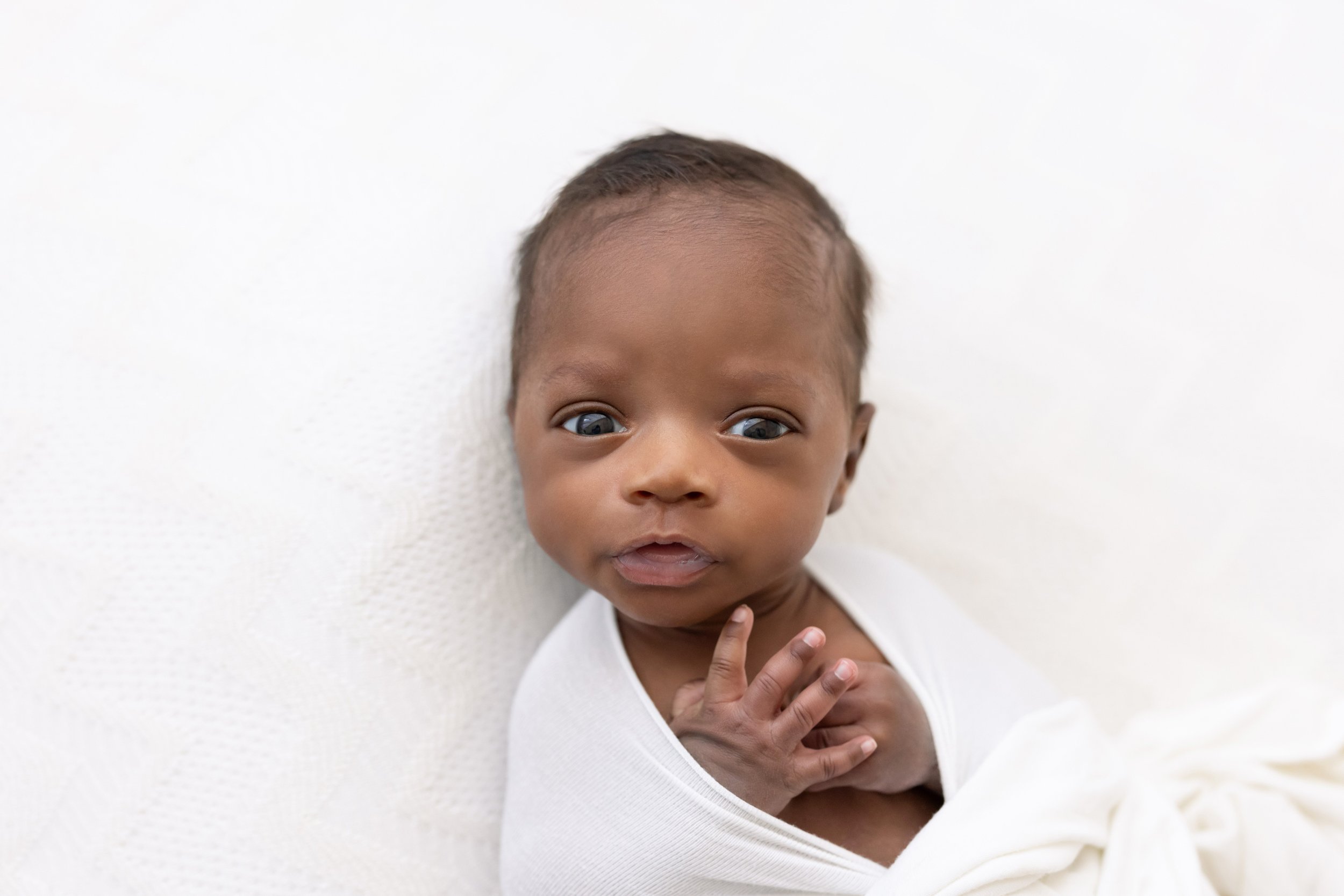  A photograph of a wide-awake African-American newborn boy with his hands sticking out of a stretchy swaddle that cradles him taken near Old Fourth Ward in a natural light studio. 