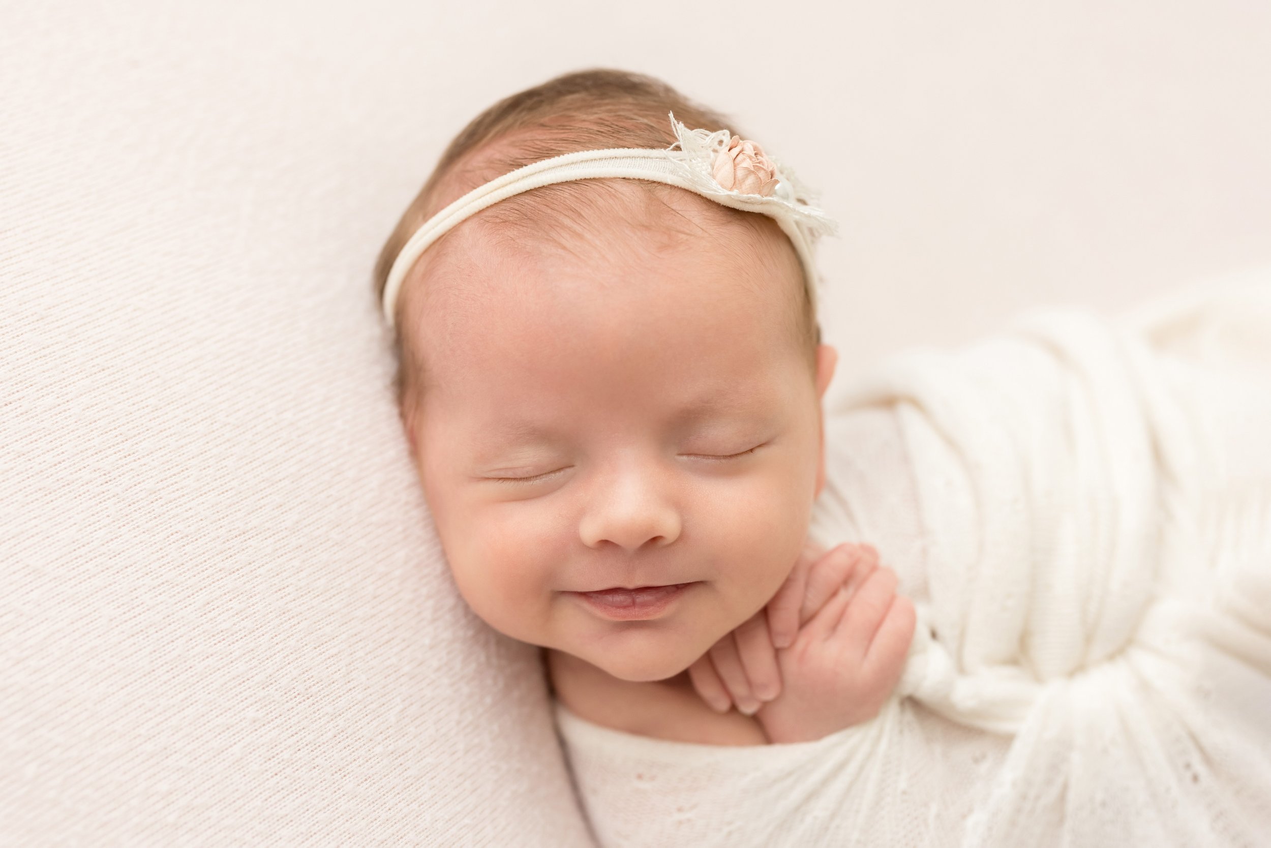  A newborn baby girl wearing a headband with a delicate rose in her wispy hair lies cradled in a stretchy swaddle with her tiny hands sticking out as she smiles in her sleep taken by Lily Sophia Photography in a studio in Ponce City Market in Atlanta