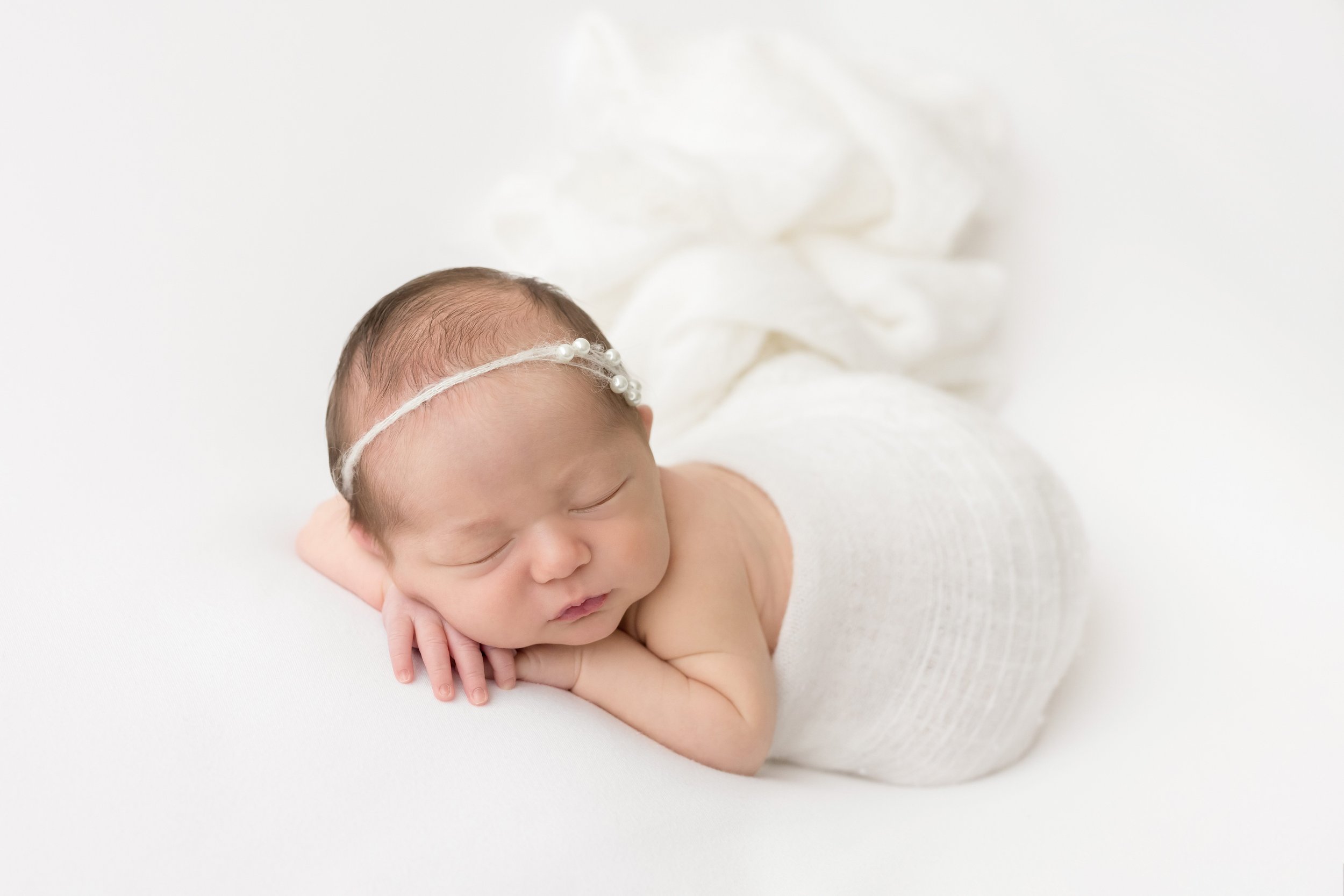  Newborn photograph of a sleeping infant girl as she lies on her stomach with her hands under her chin wearing a delicate headband in her hair and draped with a soft flowing blanket taken in a natural light studio near Smyrna in Atlanta, Georgia. 