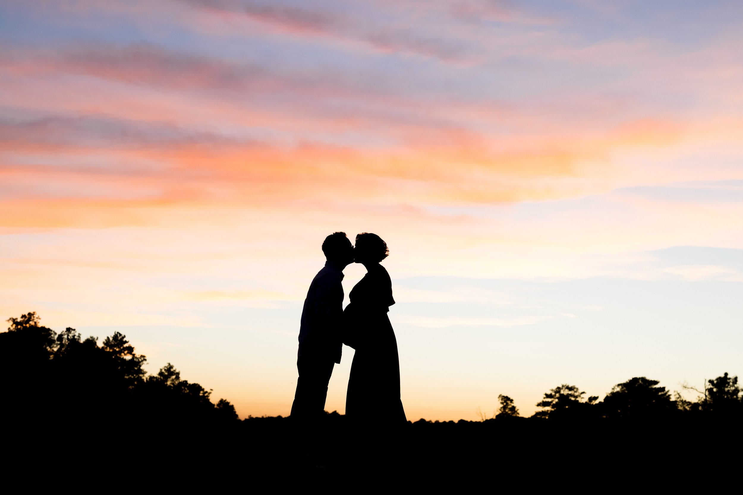  Maternity silhouetted photo of a couple as they stand in an Atlanta park at sunset and kiss one another.  