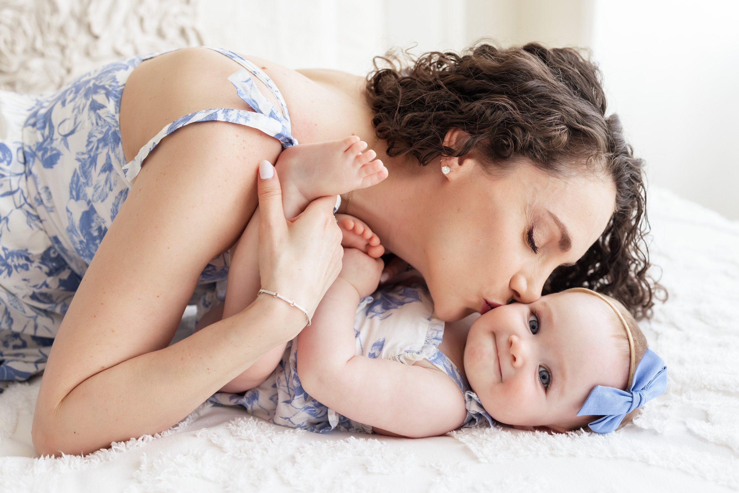  Milestone photo of a mother lying on a bed next to her young daughter who is lying on her back and kissing her chubby cheek taken in a studio near Kirkwood in Atlanta using natural light. 