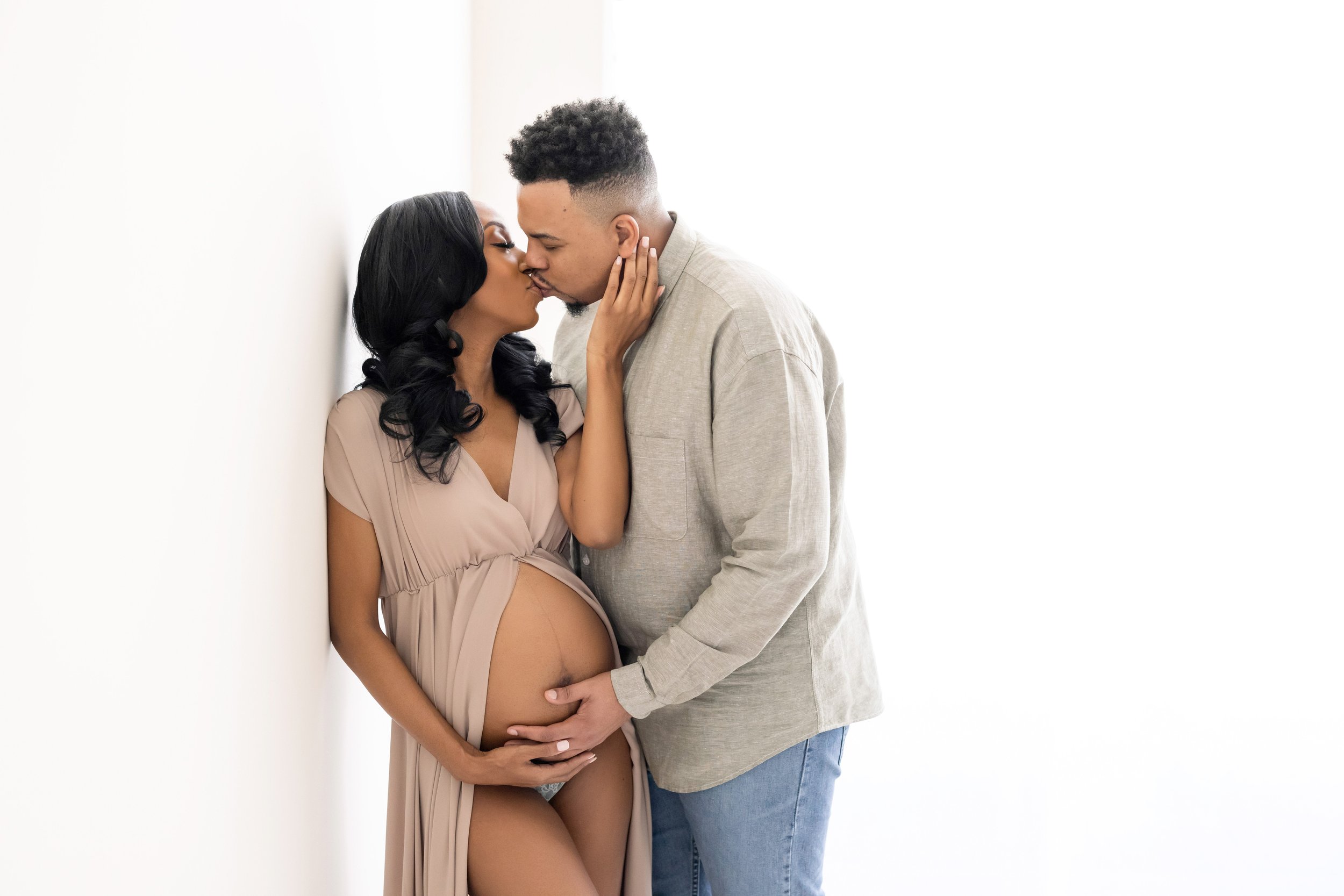  Maternity photo of an African-American couple with the pregnant woman wearing a split-front full-length gown exposing her pregnant belly as her husband stands by her side and kisses her while they place their hands on their child in utero taken in a