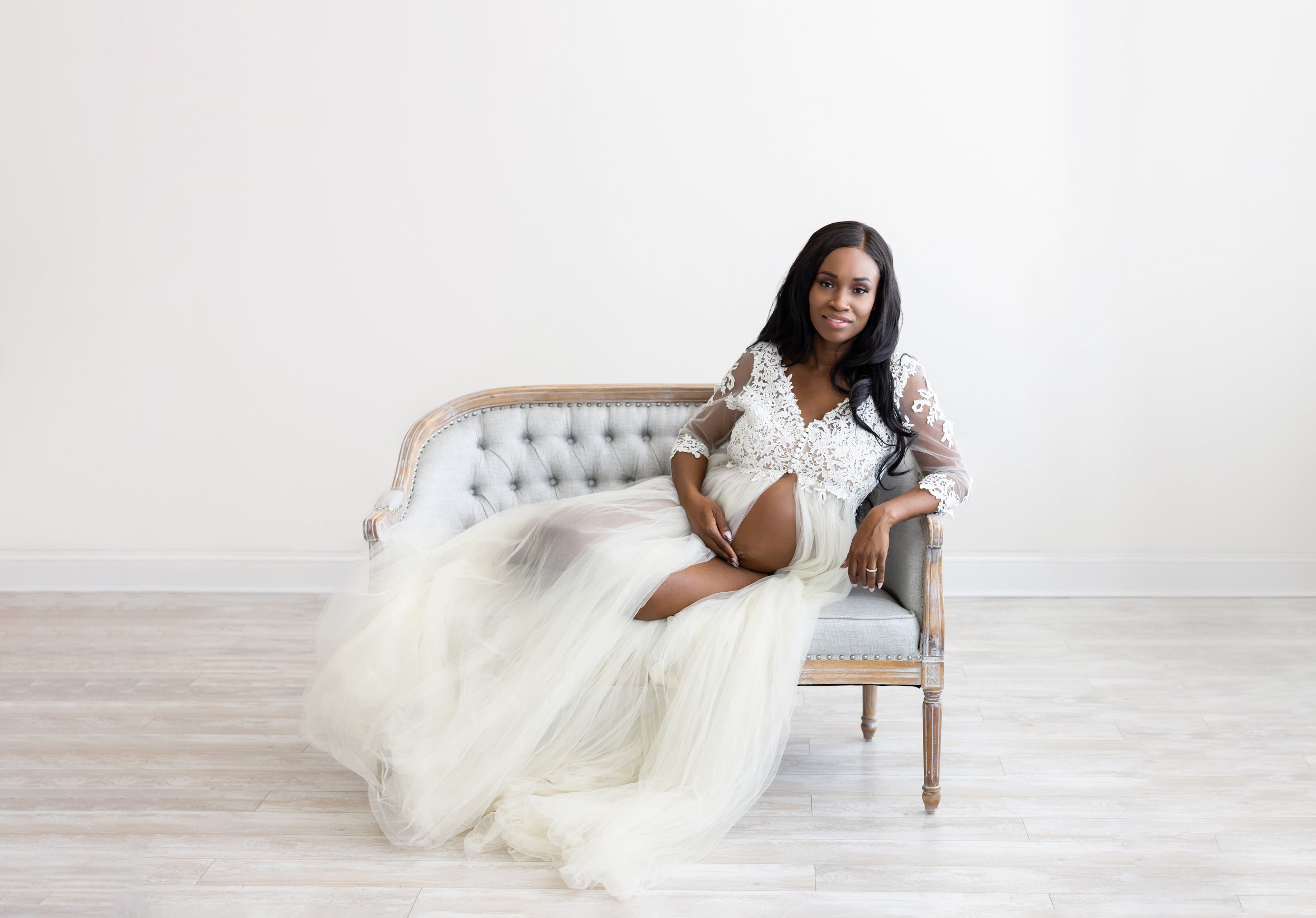  Maternity portrait of an African-American woman wearing a split front flowing full-length gown as she sits on a tufted settee leaning on her right arm and holding her bare belly taken in a studio using natural light near Buckhead in Atlanta. 