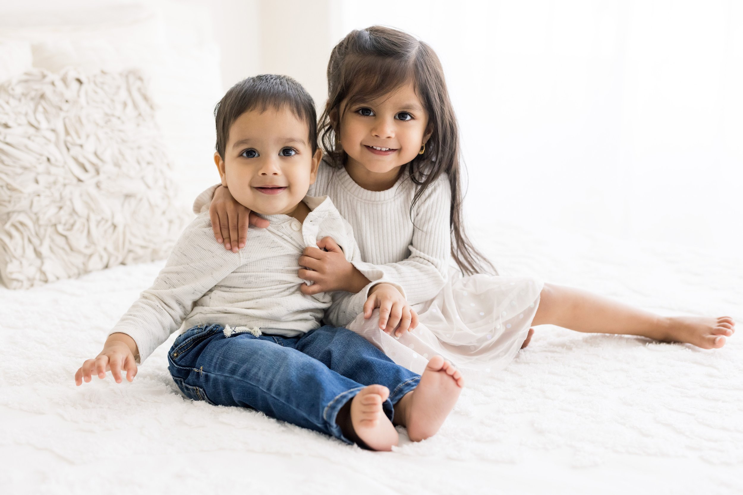  Siblings sit atop a bed with the older sister holding her baby brother as he sits with his feet in front of him in front of a window streaming natural light in a studio near Virginia Highlands in Atlanta. 