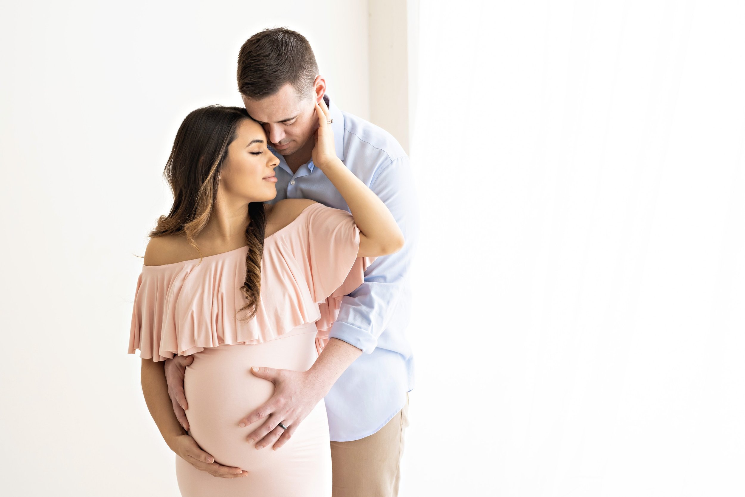  Maternity photo session wit a pregnant mother wearing an off the shoulder stretchy full-length gown with a large ruffle bodice holding her belly with her right hand and placing her left hand on the cheek of her husband as he stands behind her and pl