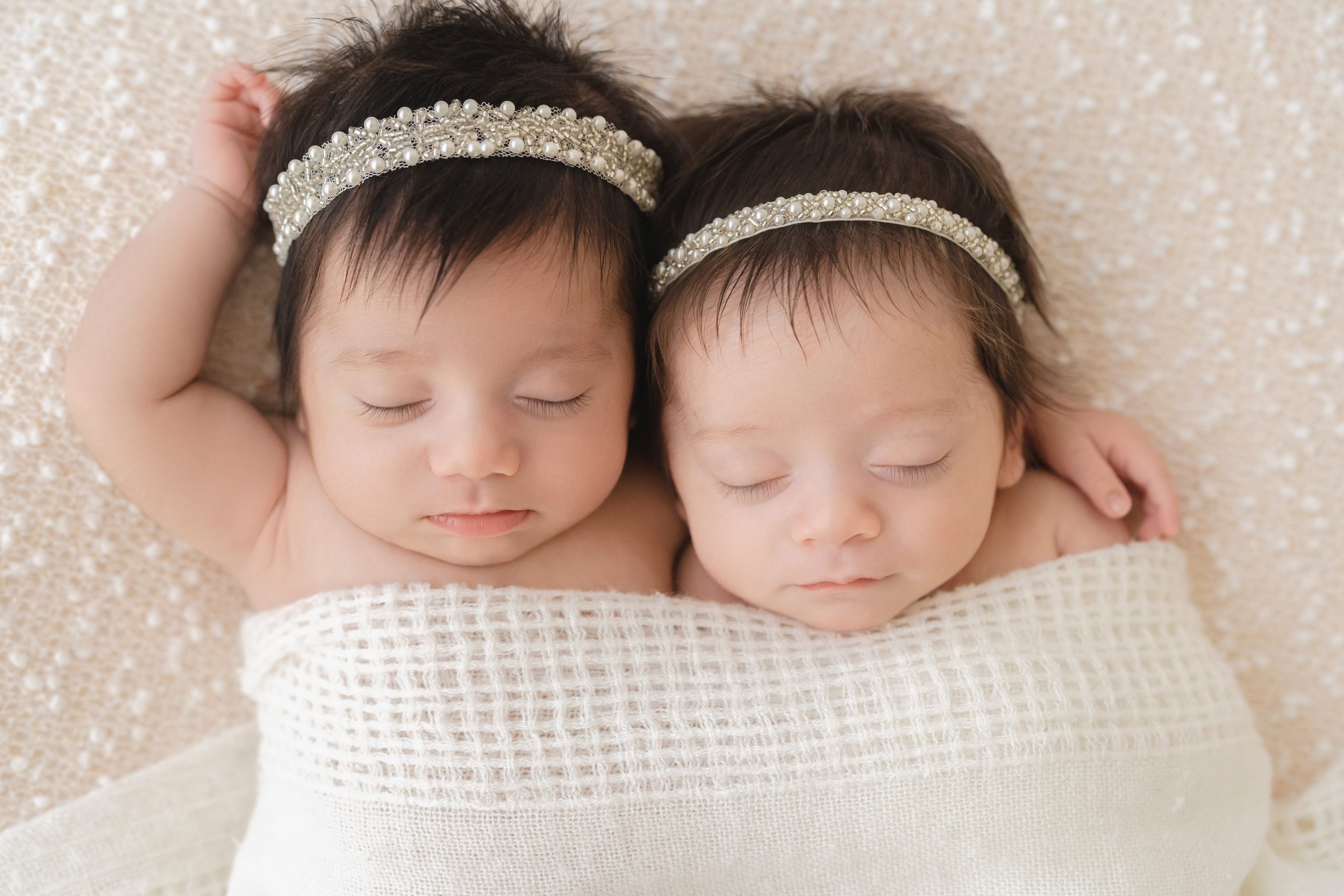  Newborn portrait of Asian twin girls as they lie on their backs on a bed with one of the twins wrapping her arm around her sister and raising her other hand in the air as the two of them are wrapped together in a blanket taken in a studio in Ponce C